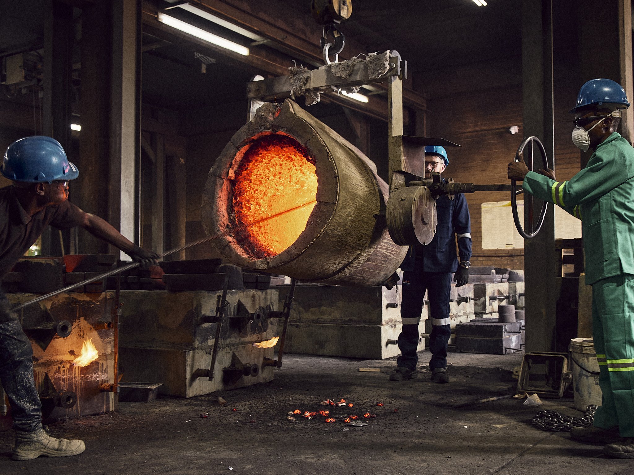 Workers in safety gear controlling a molten metal pour from a large, glowing orange pipe in an industrial foundry.