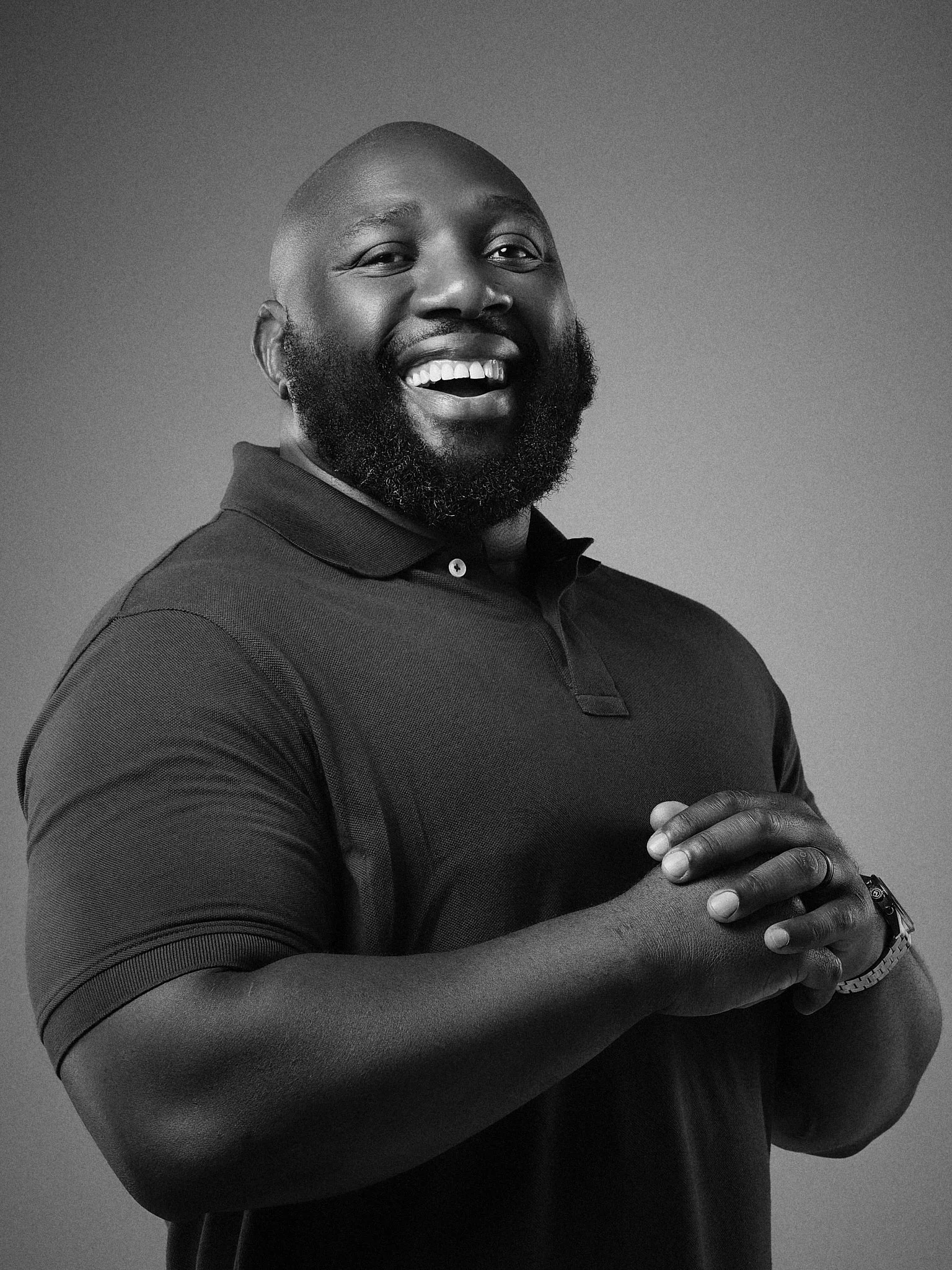 Black and white photo of a smiling man with a bald head and a beard, wearing a polo shirt, with his hands clasped together at chest level.