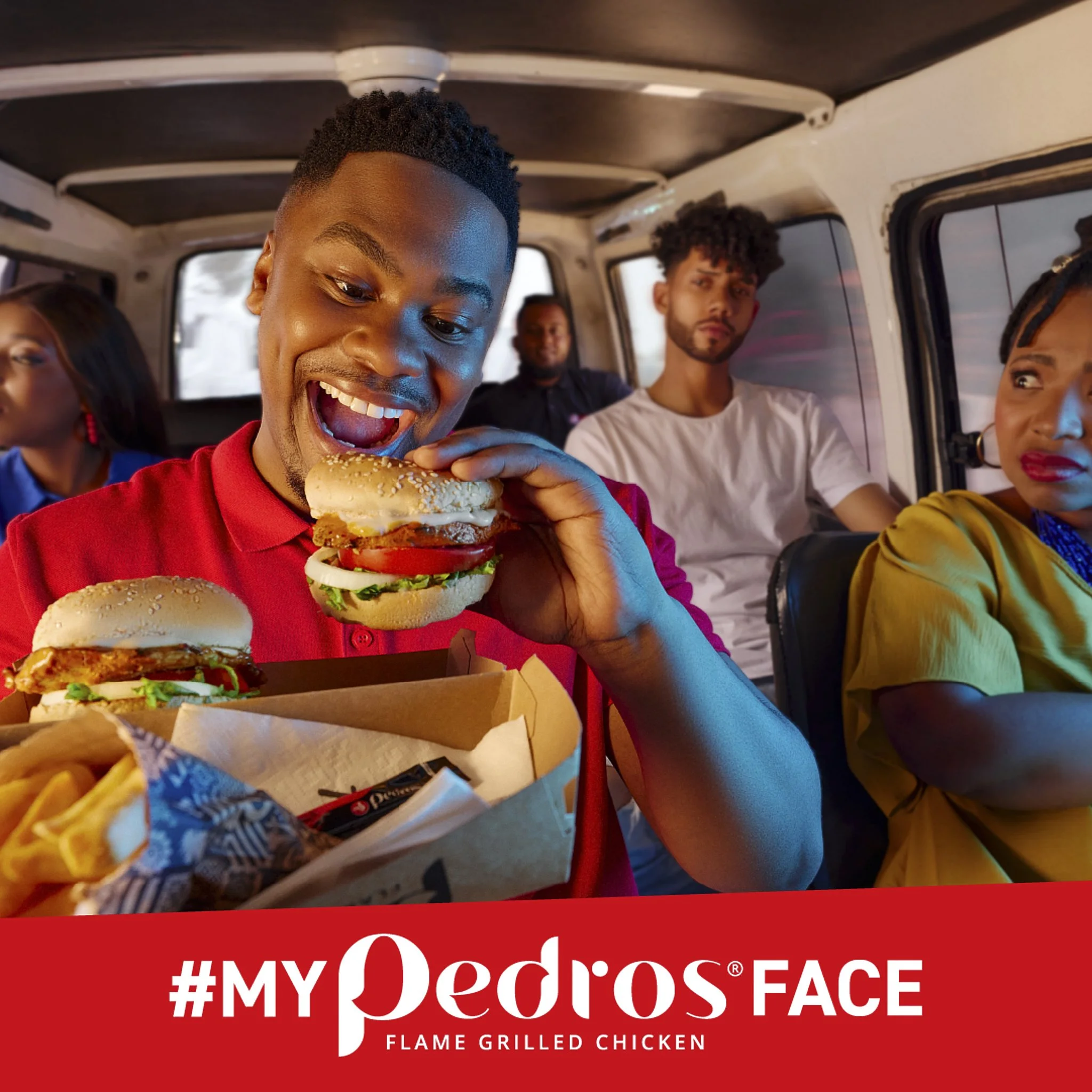 Young man with short hair smiling broadly while holding a large burger with lettuce, tomato, chicken patty, and sesame seed bun inside a vehicle. Other people sit in the background.