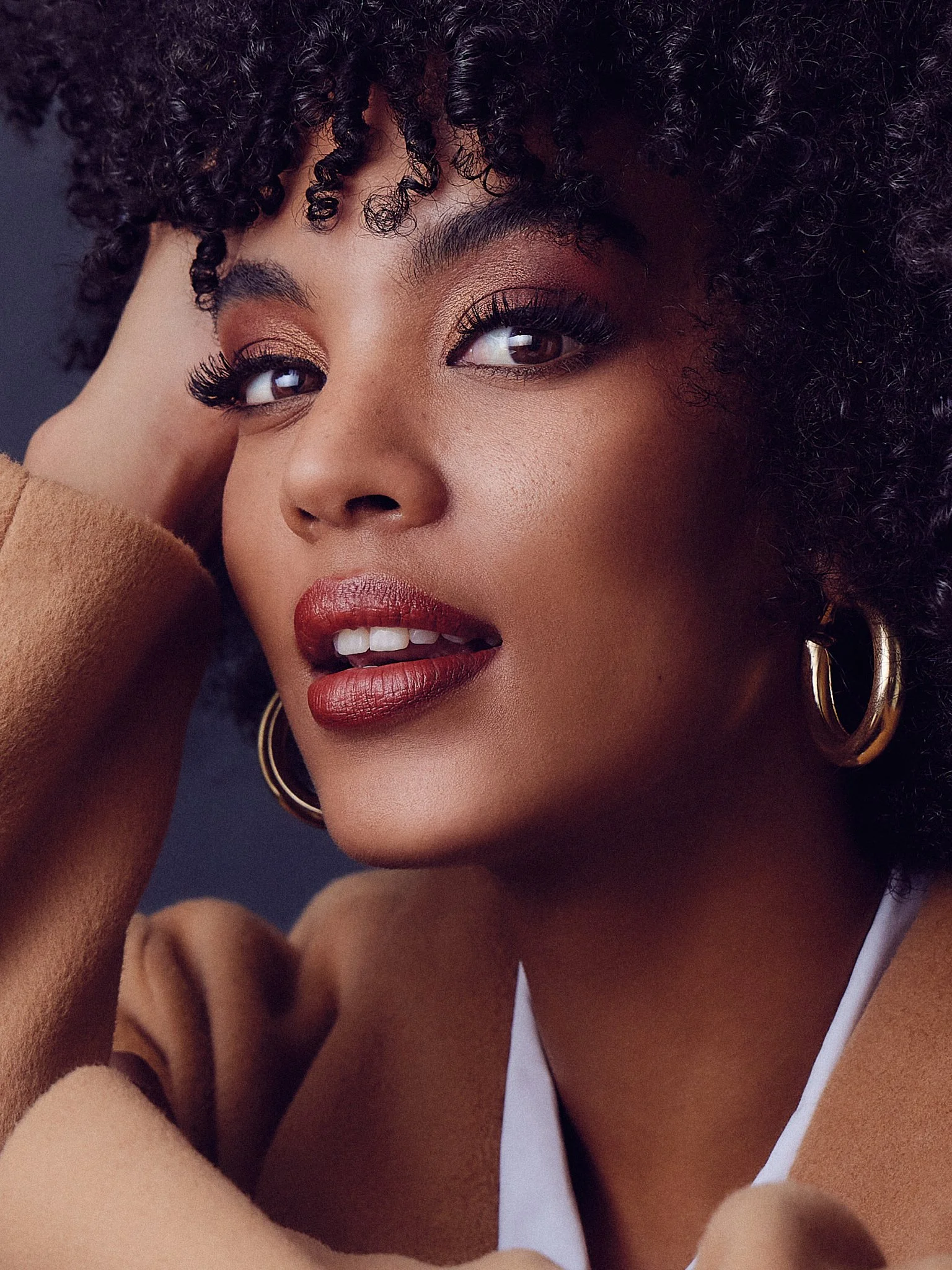 Close-up of a woman with dark, curly hair and hoop earrings, smiling with her hand near her face, wearing a beige coat and white top. Edgars winter ad