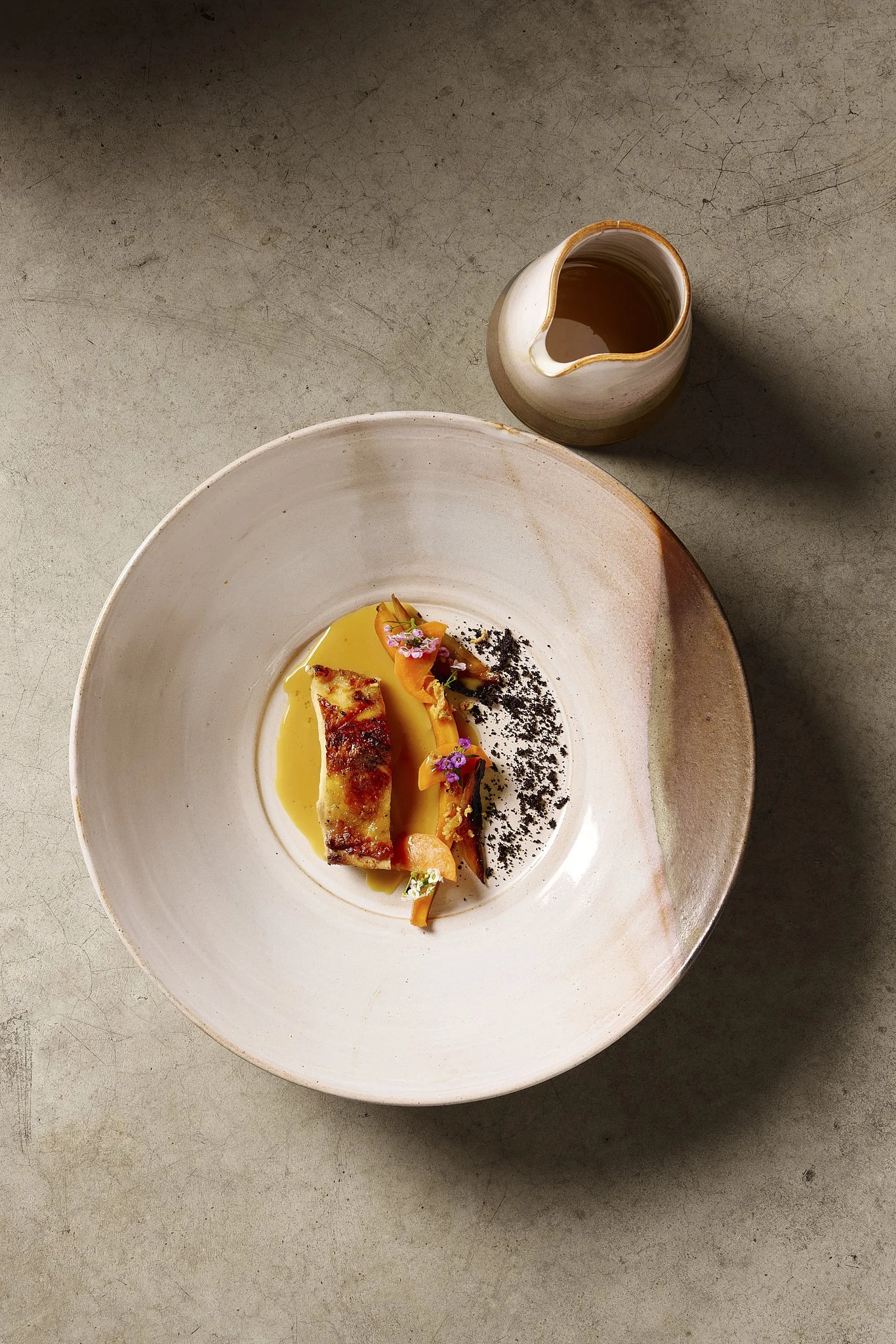 A dish with a piece of grilled fish, garnished with herbs, vegetables, and edible flowers, served with a creamy sauce in a round, white ceramic bowl. A small pitcher of sauce or broth is placed nearby on a textured gray surface.