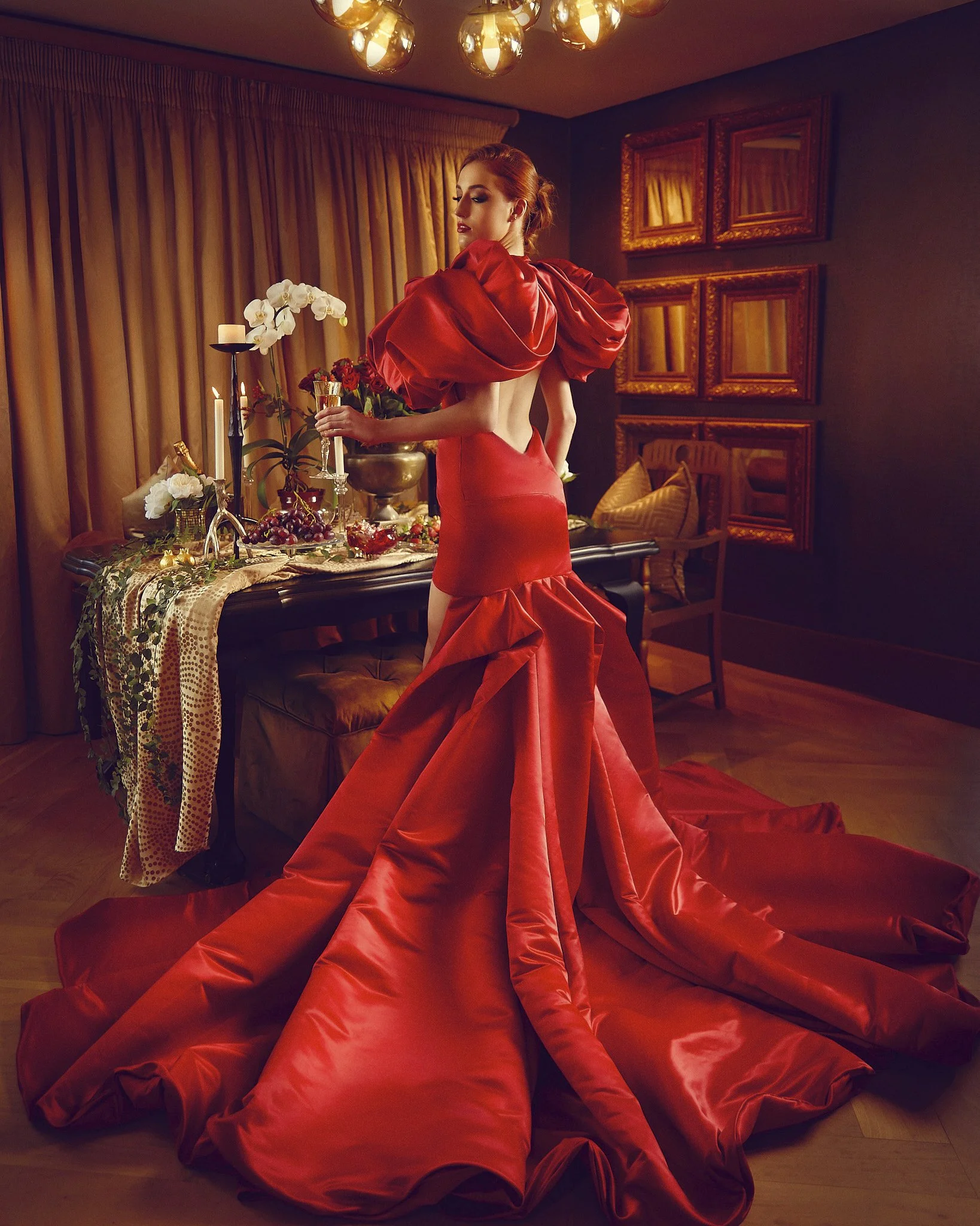 A woman in an elegant, strapless red gown with puffy sleeves stands in a warmly lit room, holding a flute glass, with a table of flowers, candles, and grapes behind her.