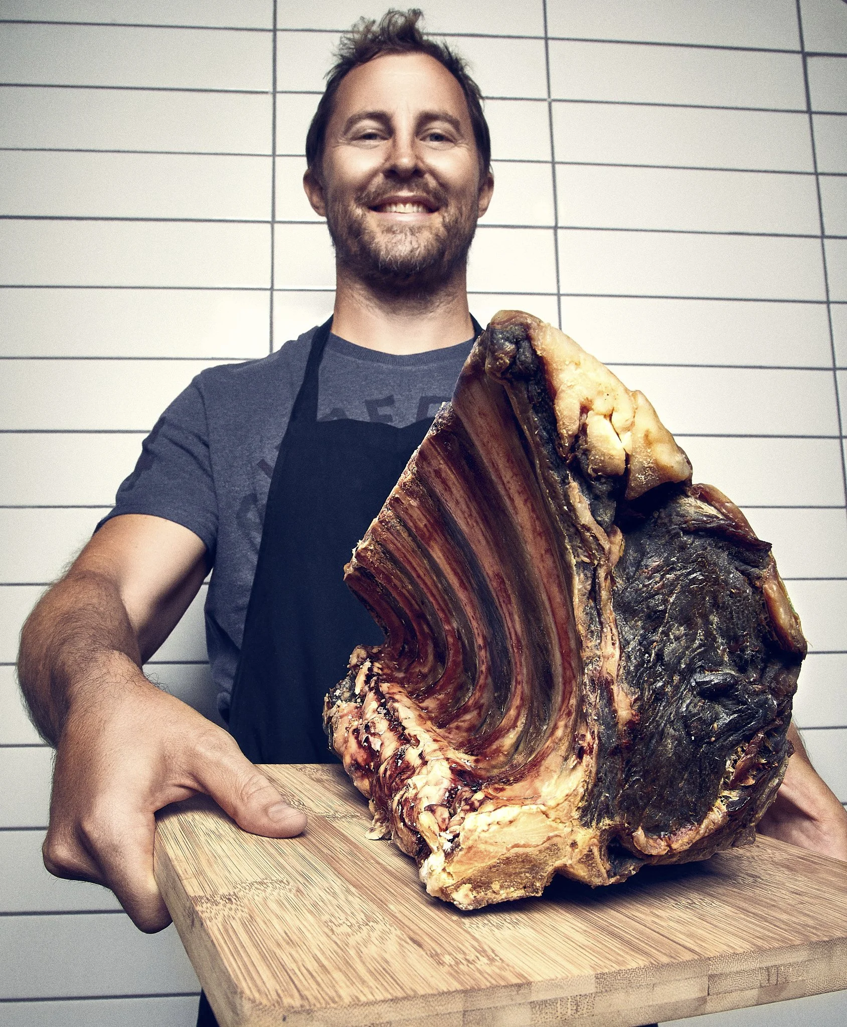 Chef Graham Nielson from 9th Ave holding a large dry aged rack of rib eye on a wooden cutting board, smiling, against a white wall with black grid lines.