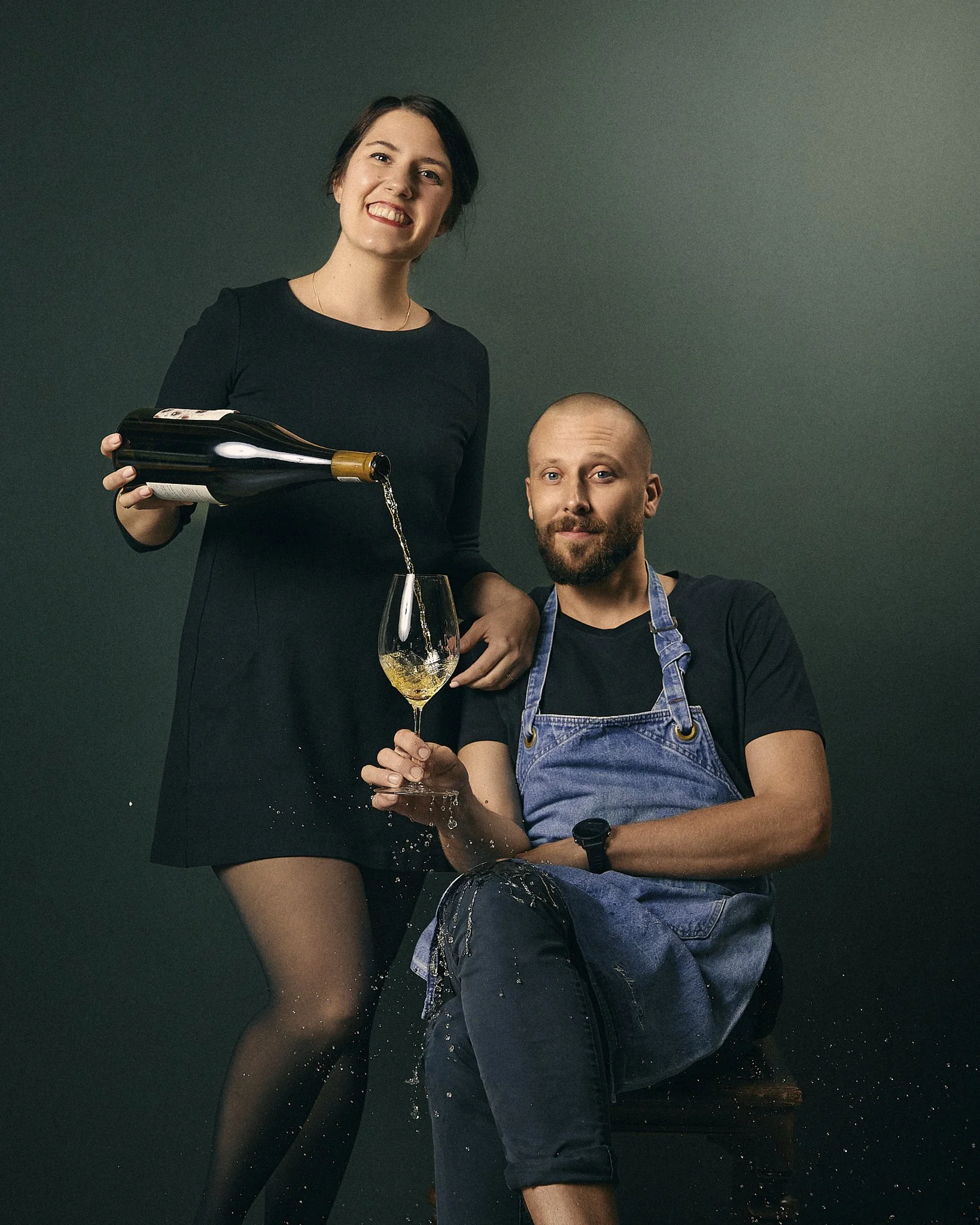 A smiling woman in a black dress pouring white wine into a glass held by a bearded man wearing a blue denim apron and black shirt. They are against a dark background, and the woman is standing while the man is sitting, with water splashing from the g