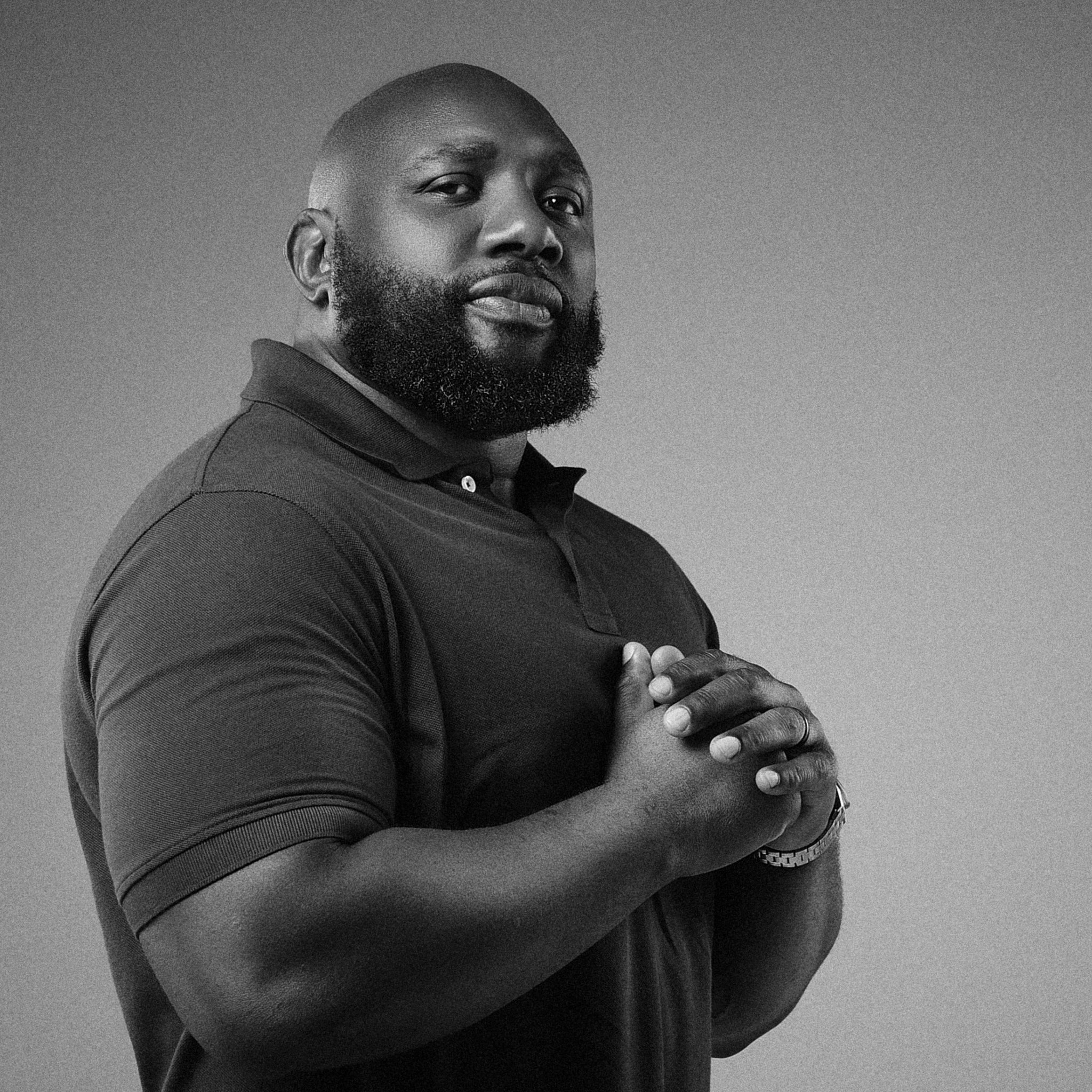 The Beast Foundation Tebdai with a beard and shaved head wearing a collared shirt, with both arms crossed, standing against a plain background.