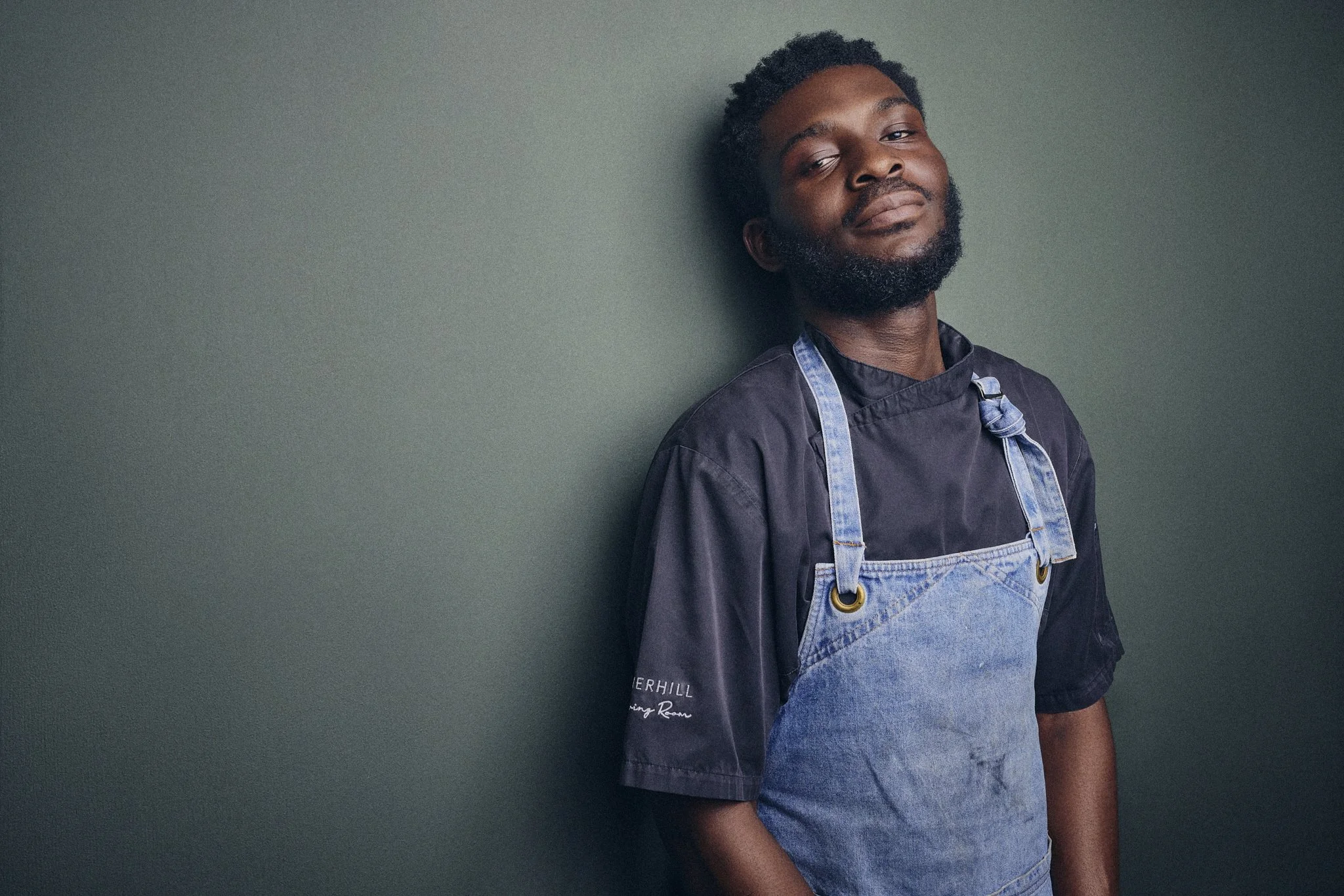 Portrait of Luxon, sous chef at The Livingroom with dark skin, short curly hair, and a beard, wearing a dark chef's uniform and denim apron, standing against a plain gray-green wall.