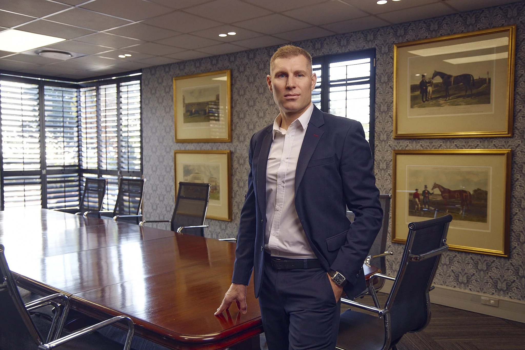 A man in a business suit standing in a conference room with large windows, framed artwork on the wall, and a wooden conference table with empty chairs.