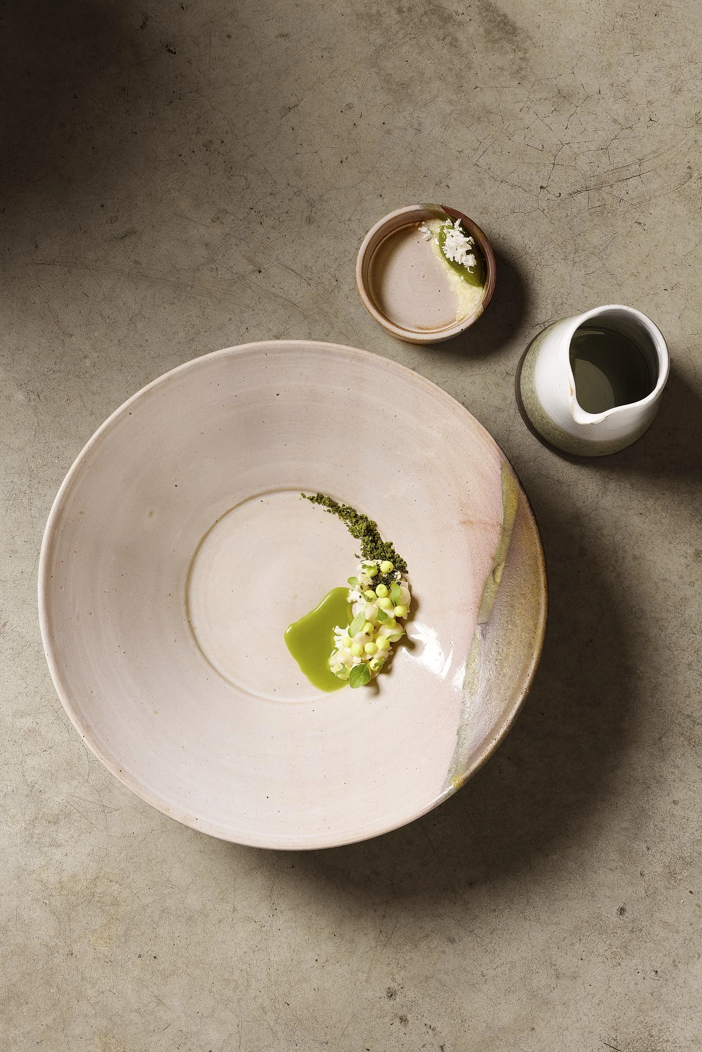 Empty pink ceramic plate with green sauce and white flowers, small jar with green liquid, and small bowl with white cream and green garnish on a gray textured surface.