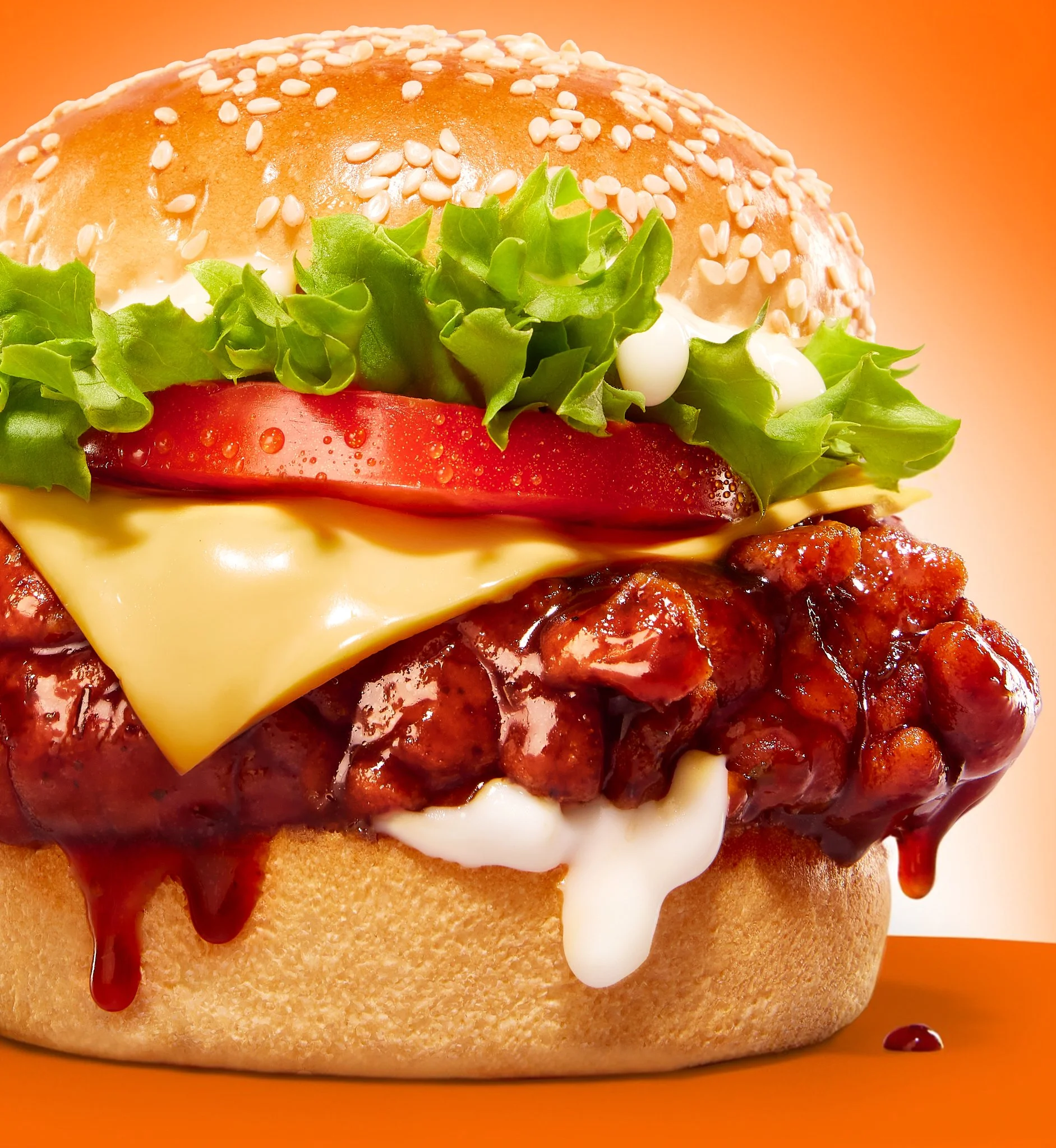Close-up of a Wacky's Dunked burger bun topped with sesame seeds, lettuce, sliced tomato, cheese, and ketchup, on an orange background.