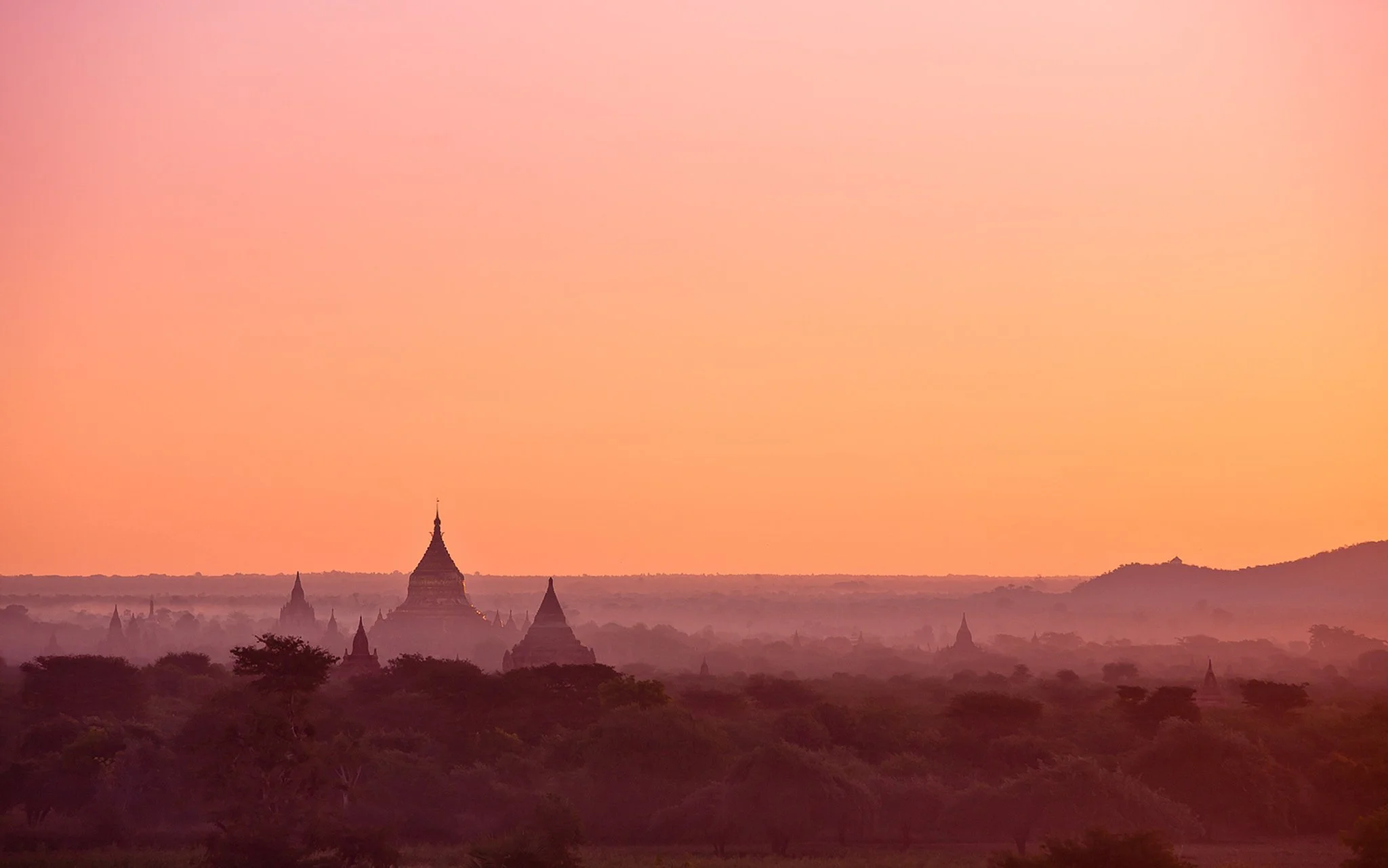 Sunset over ancient temples and pagodas in a misty landscape with trees in the foreground. The Plains of Bagan at dawn.