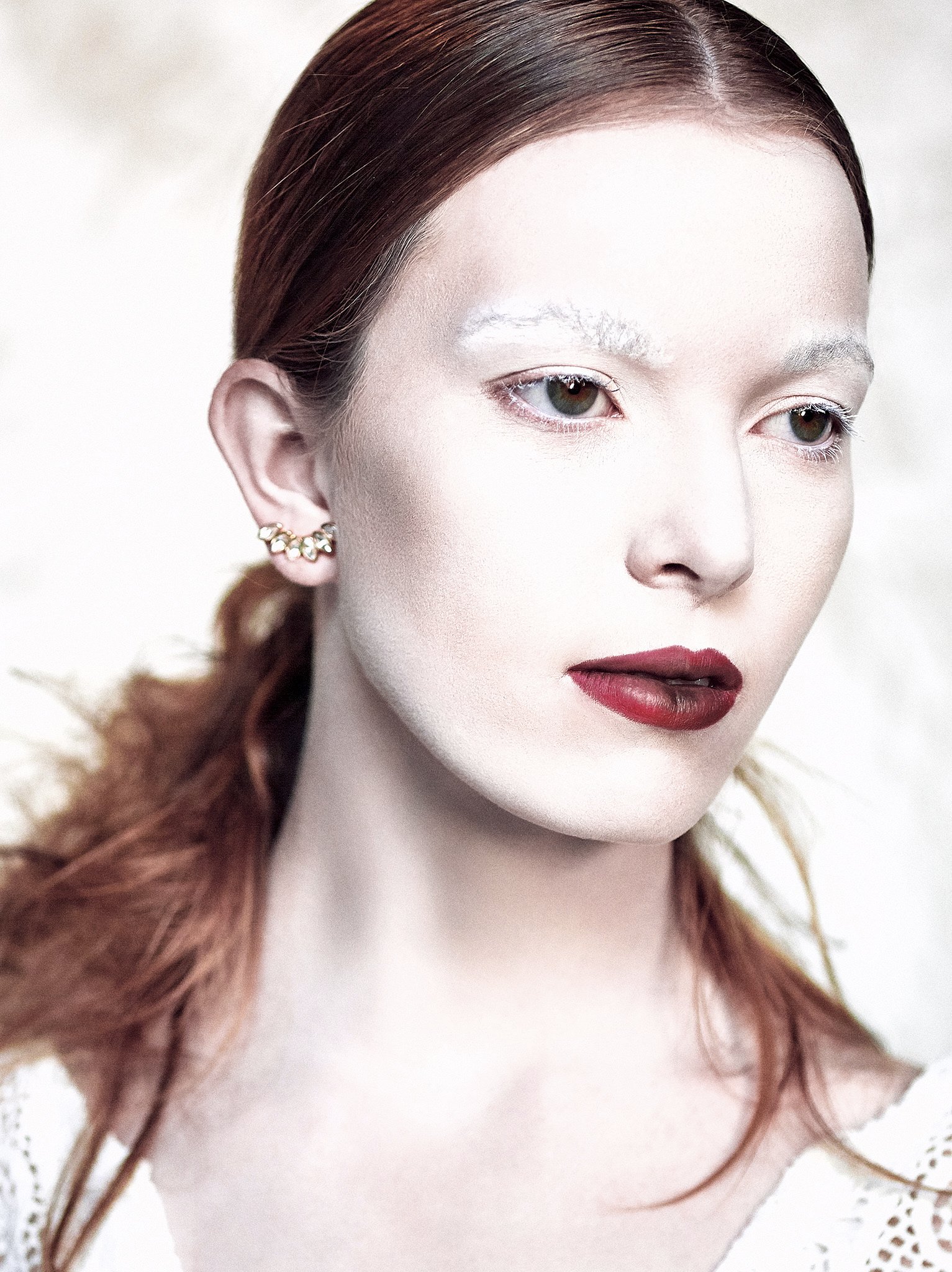 A woman with pale skin and red hair styled in loose waves. She has makeup with dark red lipstick, minimal eye makeup, and white eyebrows, wearing floral earrings. The background is light and blurred.