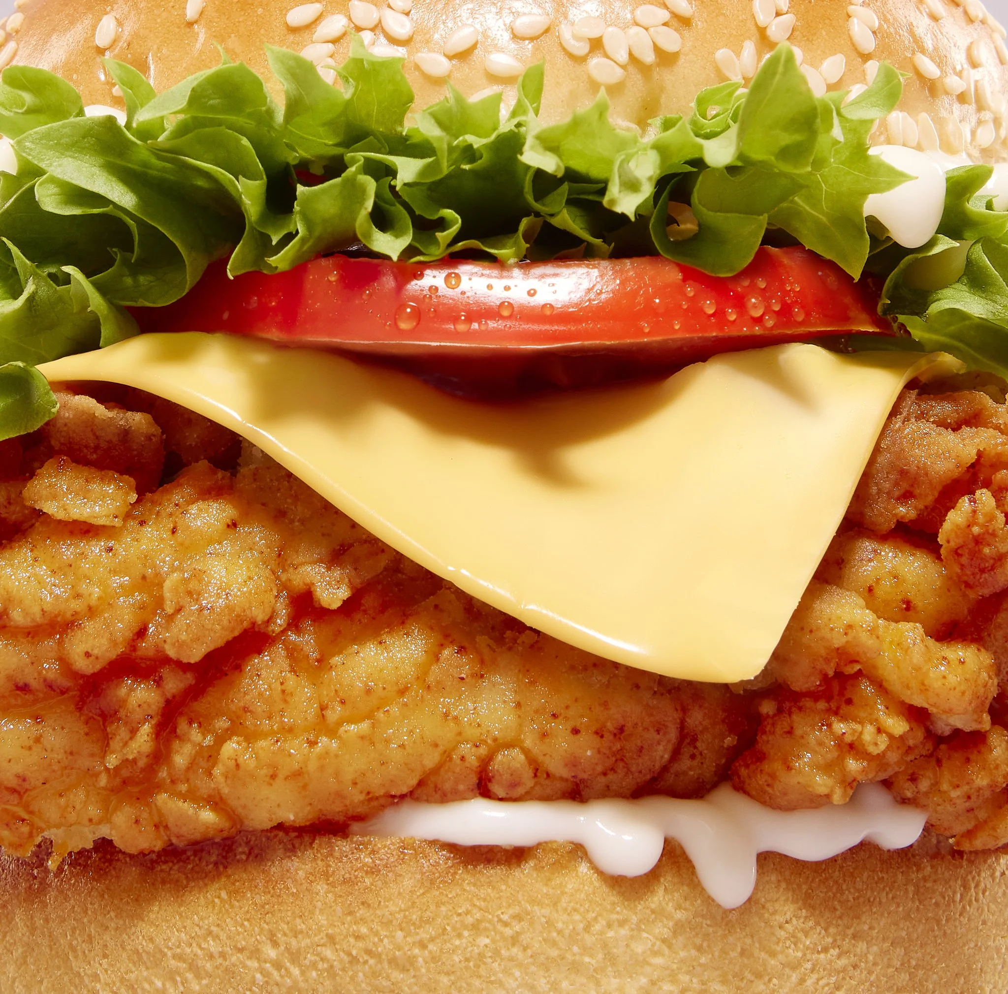 Close-up of a fried chicken cheeseburger with fried chicken, lettuce, tomato, cheese, and mayonnaise on a sesame seed bun.