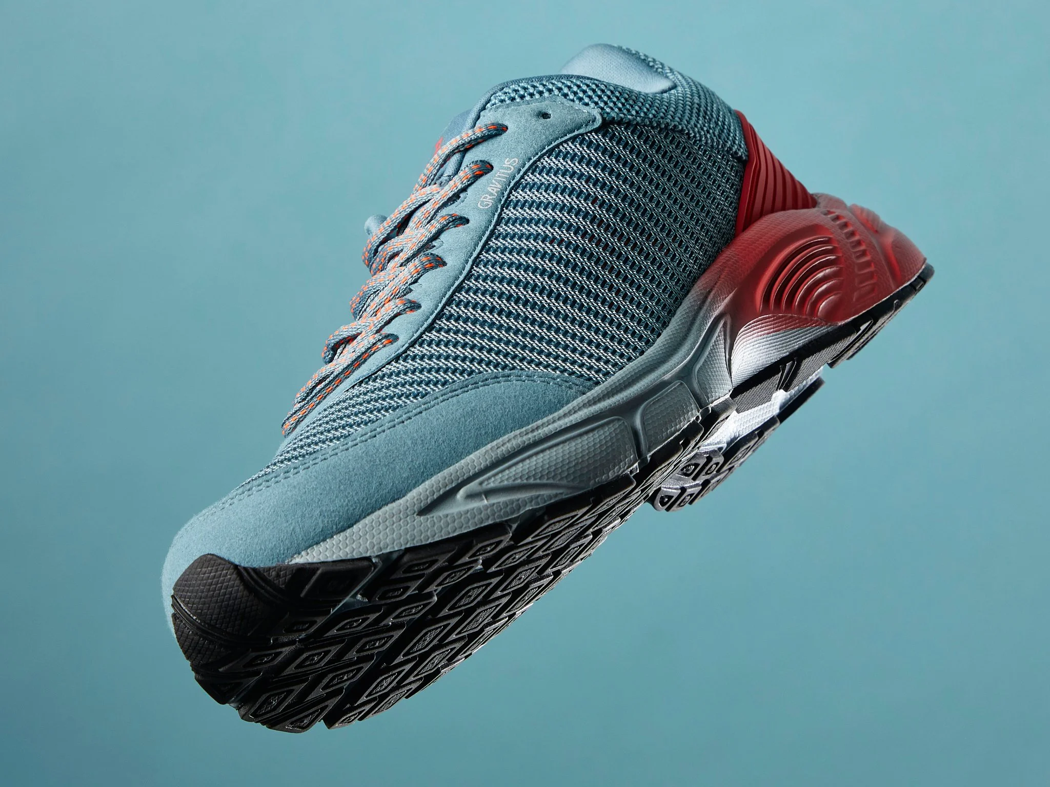 A modern athletic running shoe with a blue and grey knitted upper, red accents on the sole, and black and white patterned outsole, photographed against a light blue background.