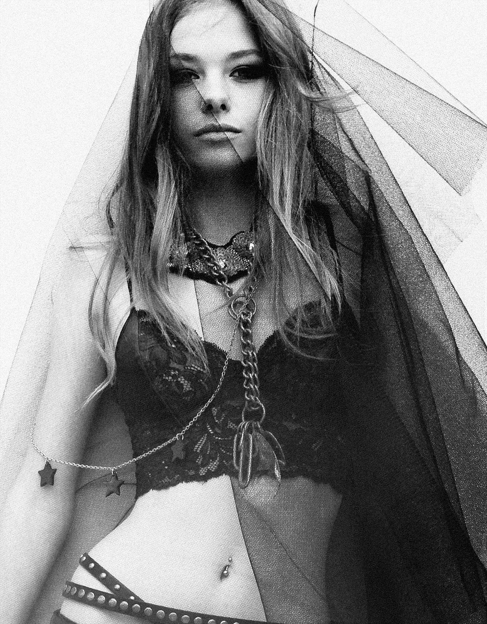 Black and white portrait of a woman with long hair, wearing a veil, layered necklaces, a leather top, and star-shaped earrings, with decorative elements around her neck and shoulders.