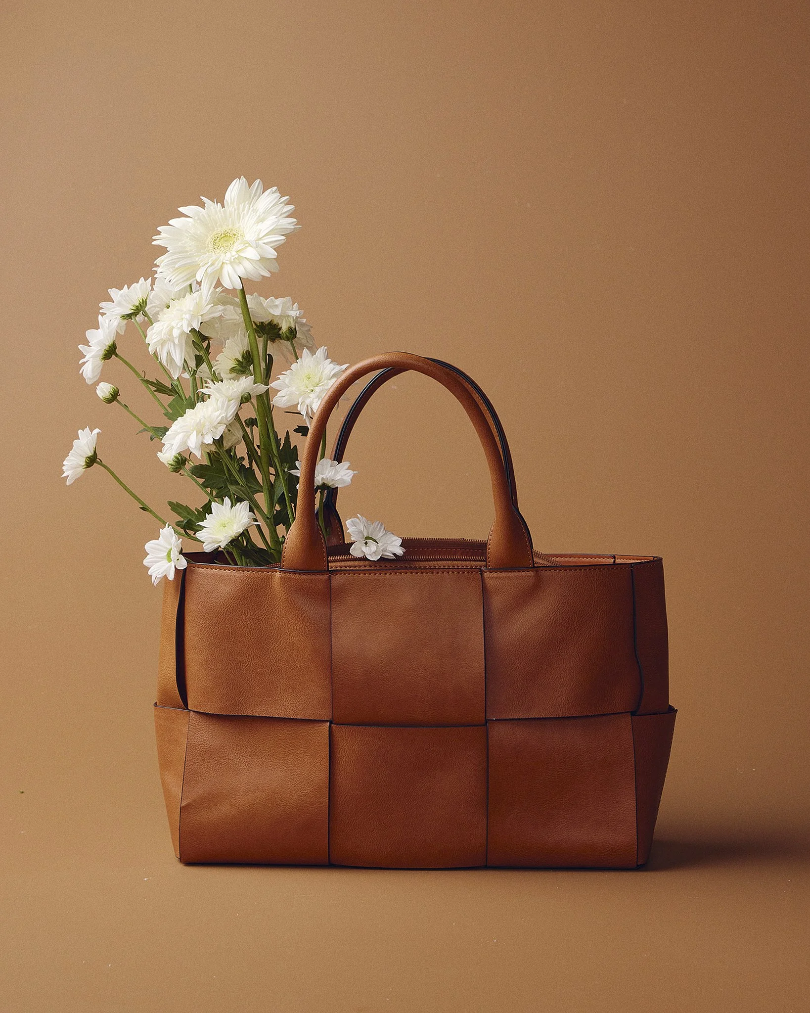 A brown leather handbag with a floral arrangement of white daisies inside, set against a beige background.