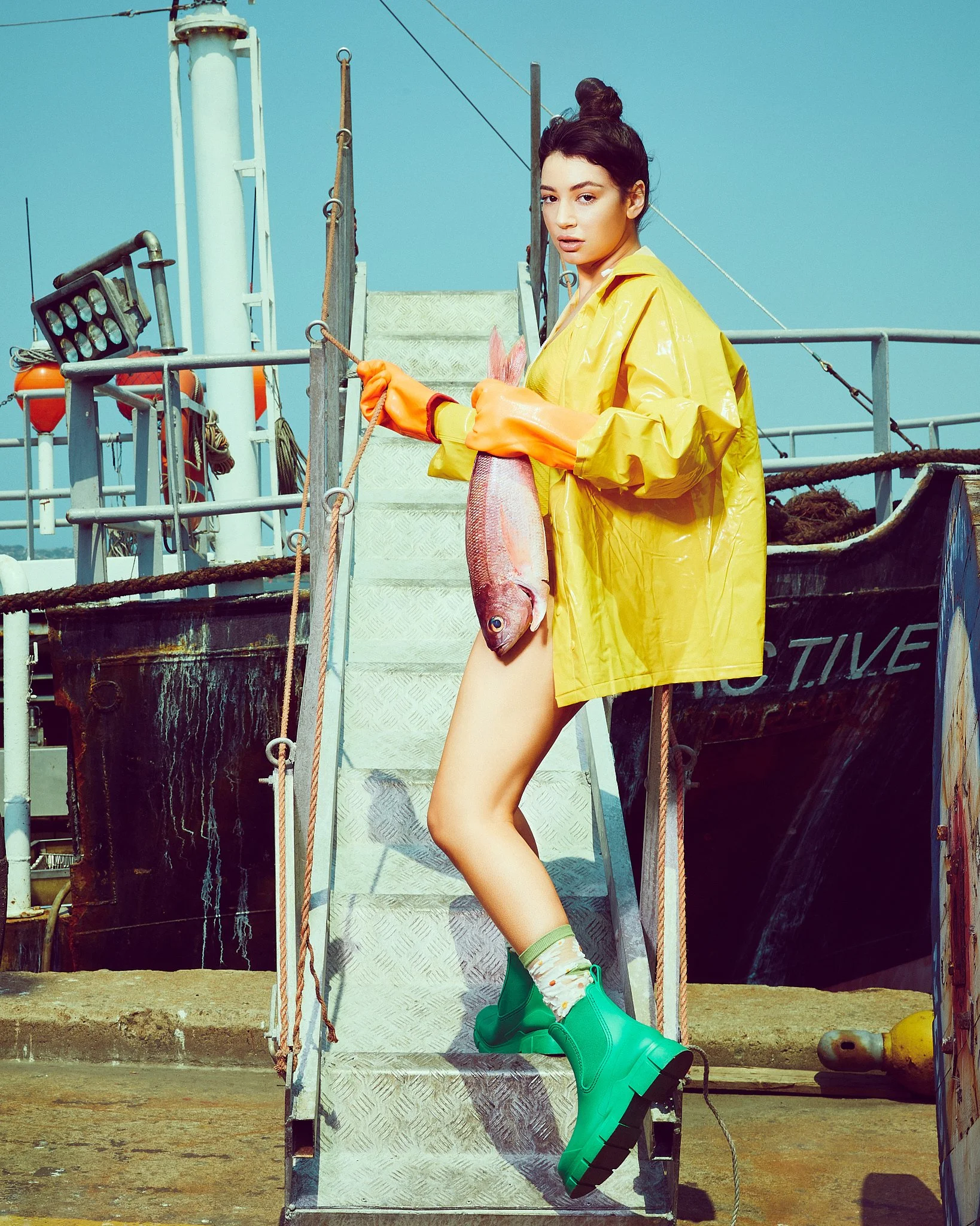 A woman standing on the stairs of a boat holding a fish, wearing a yellow raincoat, colorful socks, and green rain boots, with a clear blue sky in the background.