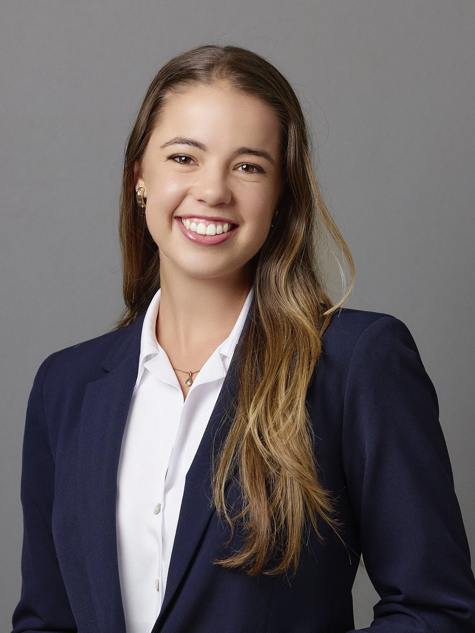 A young woman with long wavy hair wearing a navy blazer over a white shirt, smiling, against a neutral grey background.