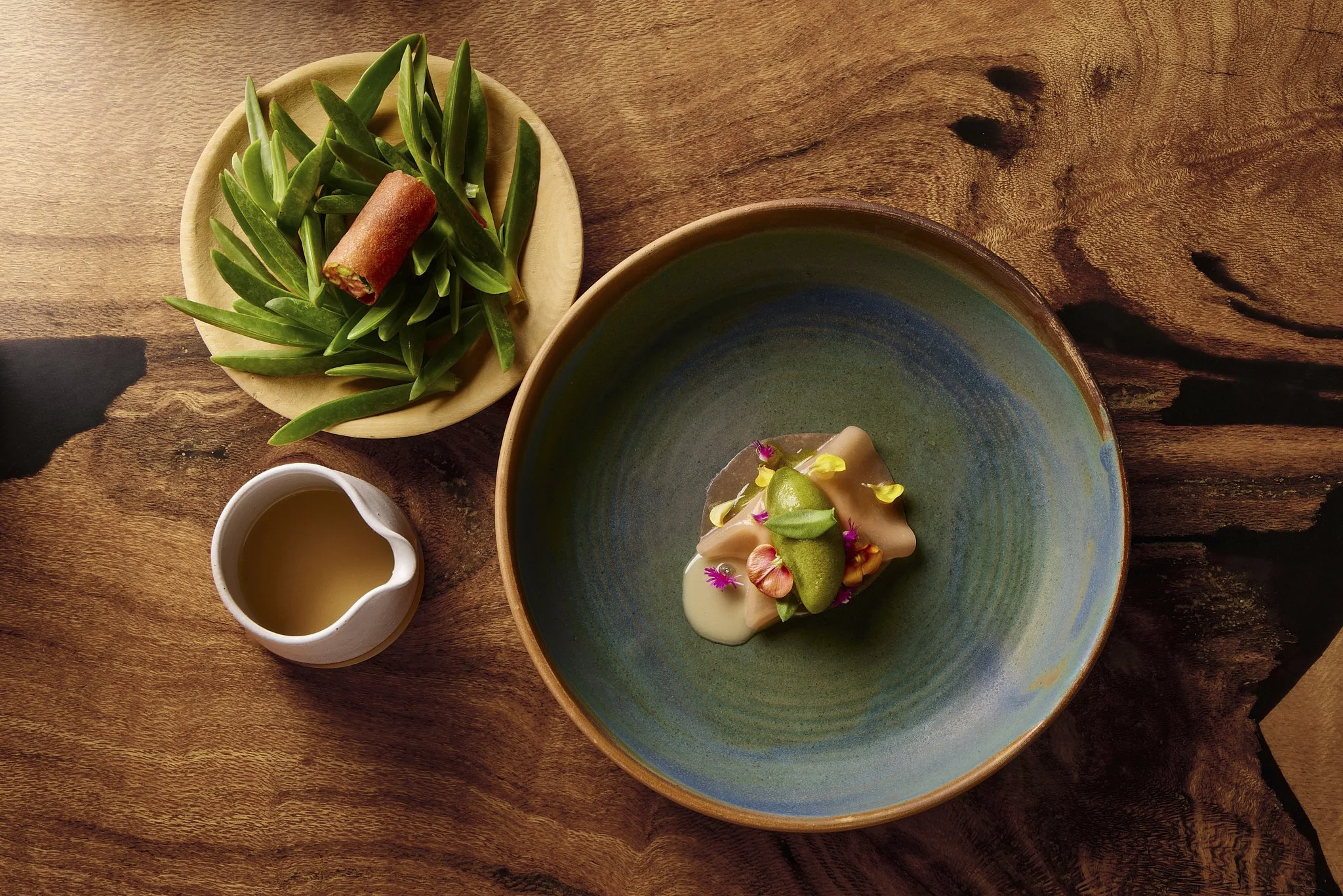 A wooden table with a green ceramic plate holding a dessert garnished with edible flowers, a small white pitcher with a sauce, a round wooden bowl filled with green succulent plants, and a small white jug also filled with a liquid.