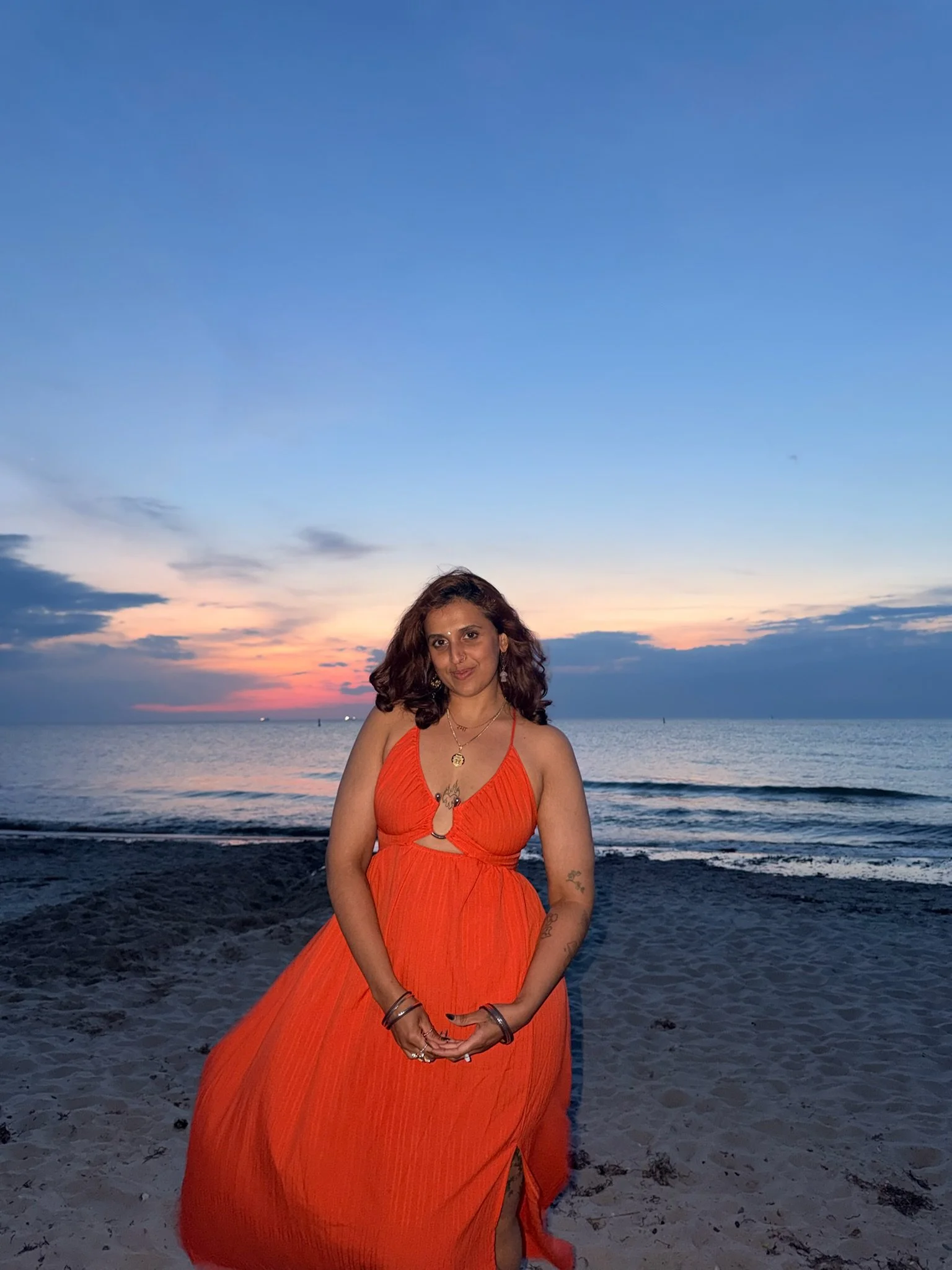 Rma, a genderfluid marathi femme, in an orange dress standing on a beach at sunset with the ocean behind them.