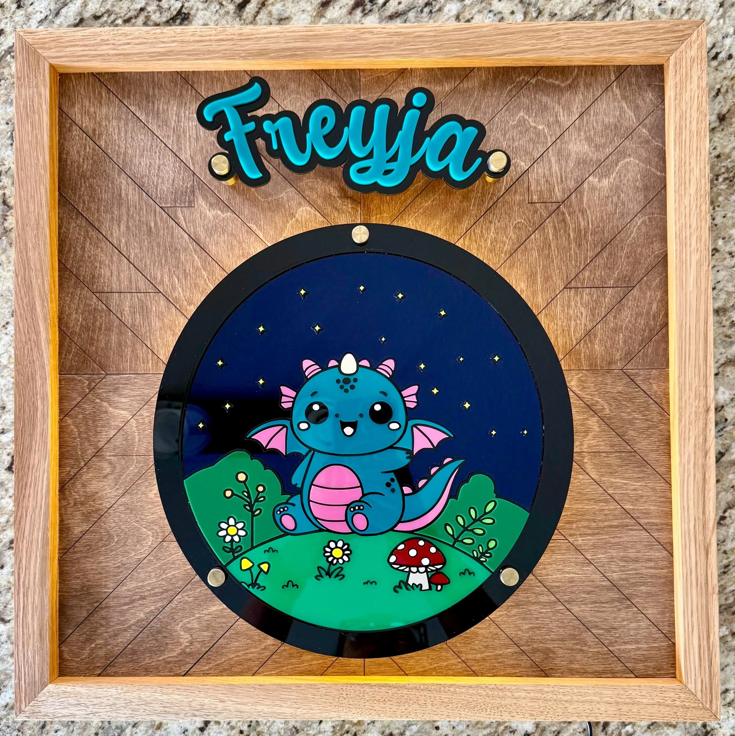 Decorative wall art featuring a cartoon blue dragon with pink wings, sitting on green grass with white daisies and red mushrooms, under a starry sky, framed with wooden edges.