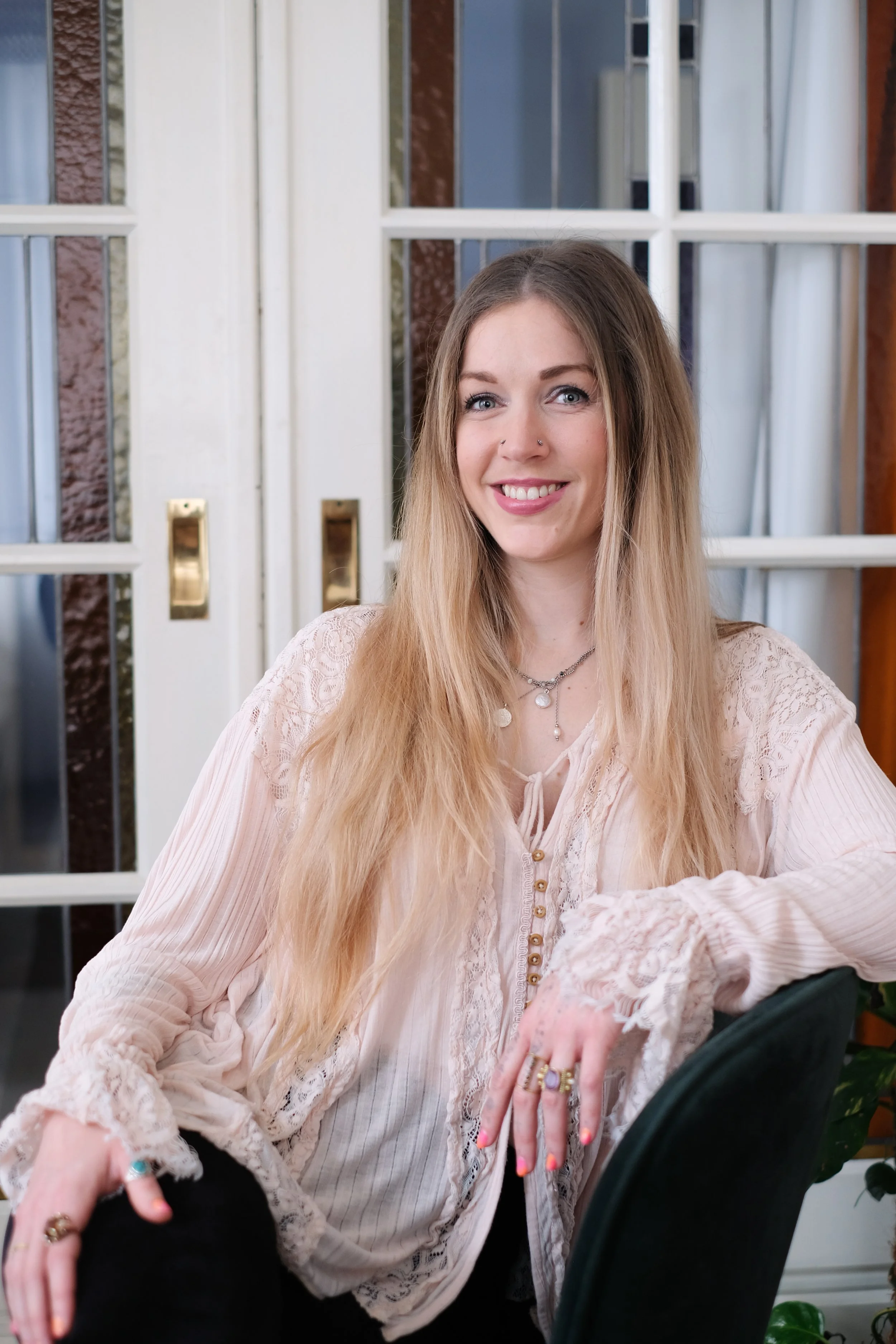 A young woman with long blonde hair, light skin, wearing a pink lace blouse, sitting at a table in front of a window with white frames. She has a nose piercings, layered necklaces, and rings on her fingers, smiling and looking at the camera.