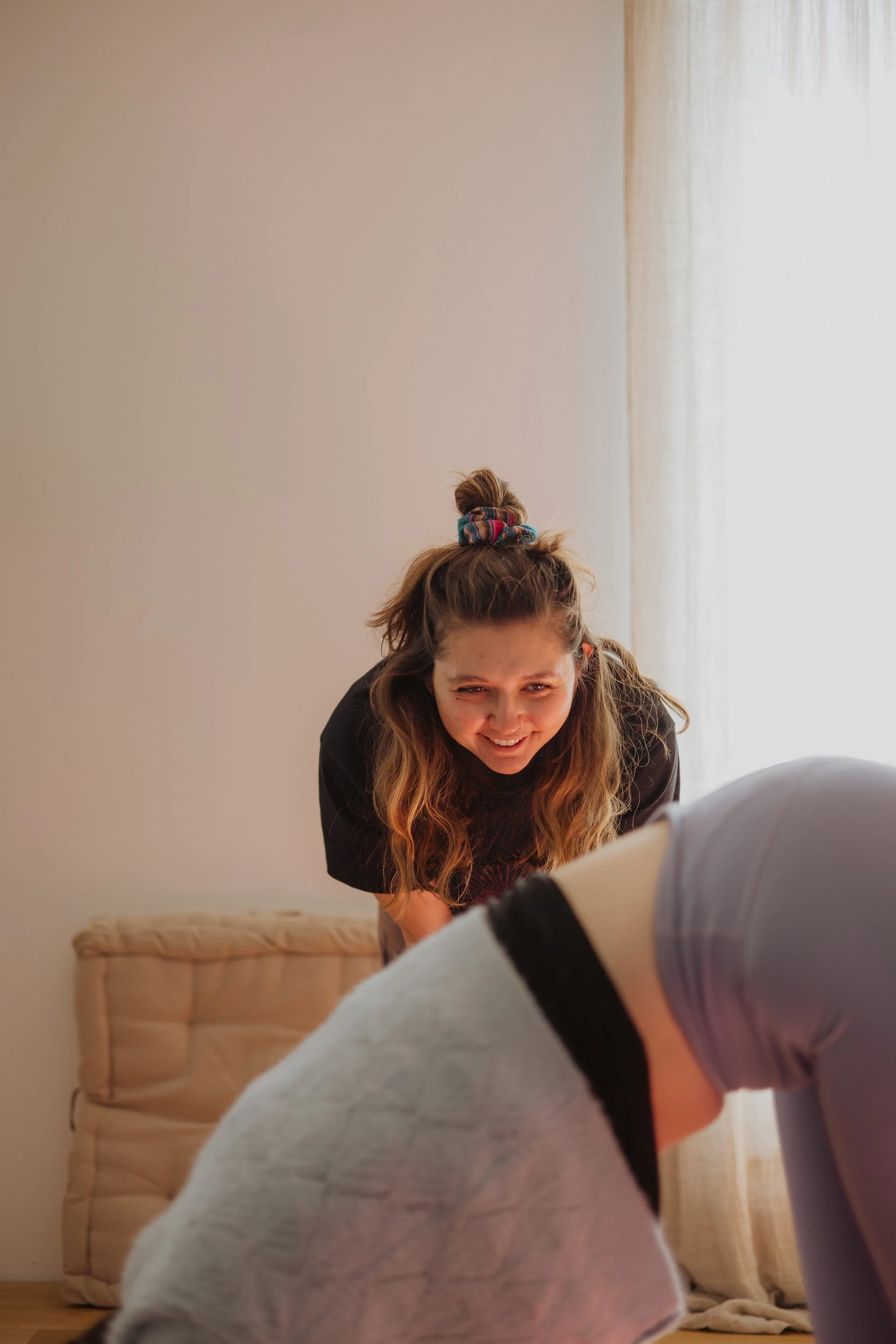 Une femme souriante, avec des cheveux attachés en bun avec un foulard coloré, est inclinée vers une élève dont on ne voit que la tête et une épaule. Elle l'ajuste dans la position de yoga du chien tête en bas.