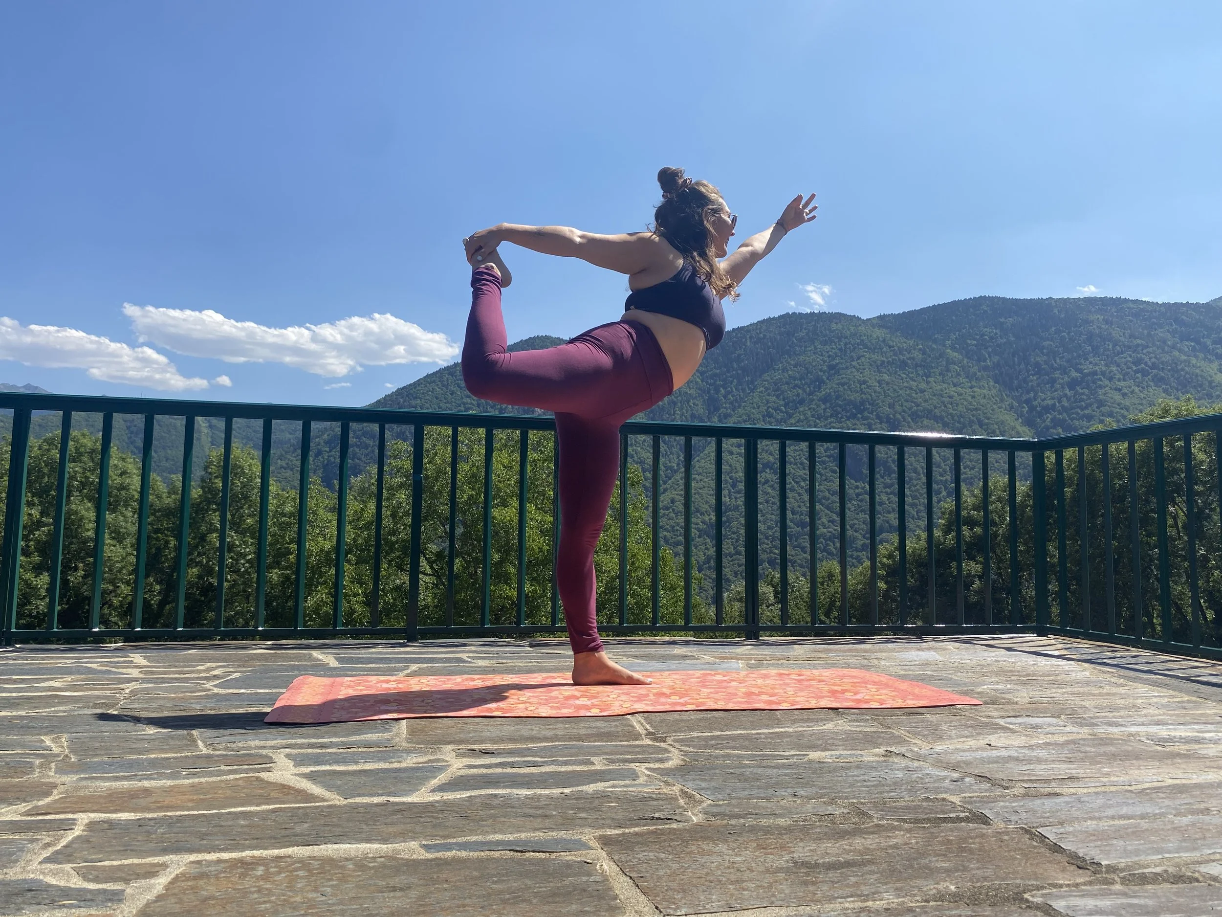 Une femme pratique le yoga en équilibre sur une jambe, en position de la danseuse, sur un tapis rose fleuri, avec un paysage de montagnes verdoyantes et un ciel bleu clair en arrière-plan.