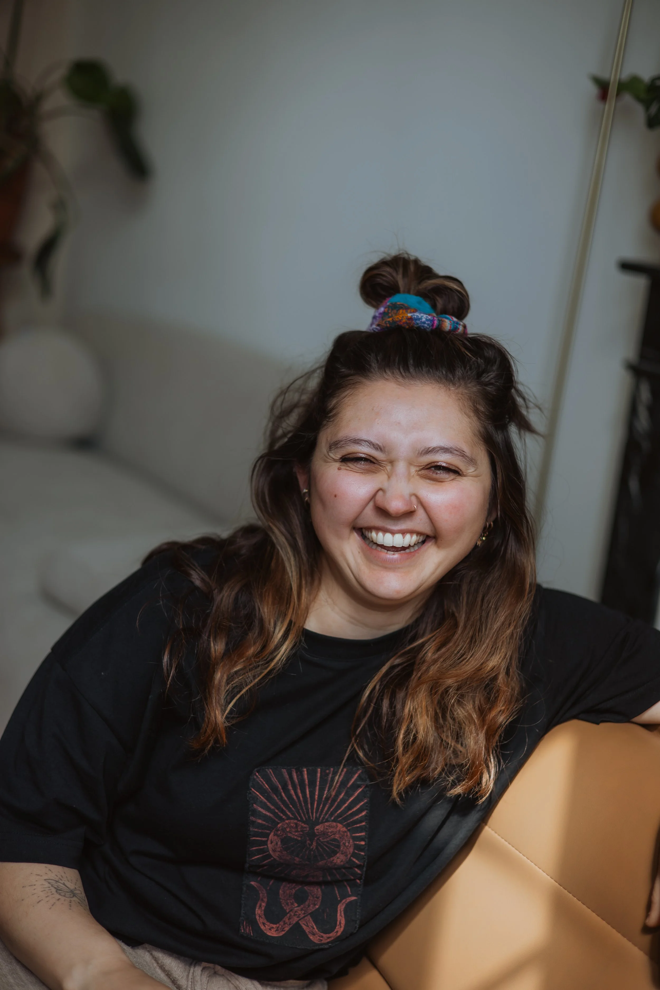 Une femme souriante avec des cheveux bruns ondulés portant un t-shirt noir avec un motif de serpent bronze et de la peau couleur crème, assise sur un canapé beige en cuir, avec un fond de mur blanc et des décorations végétales en arrière-plan.