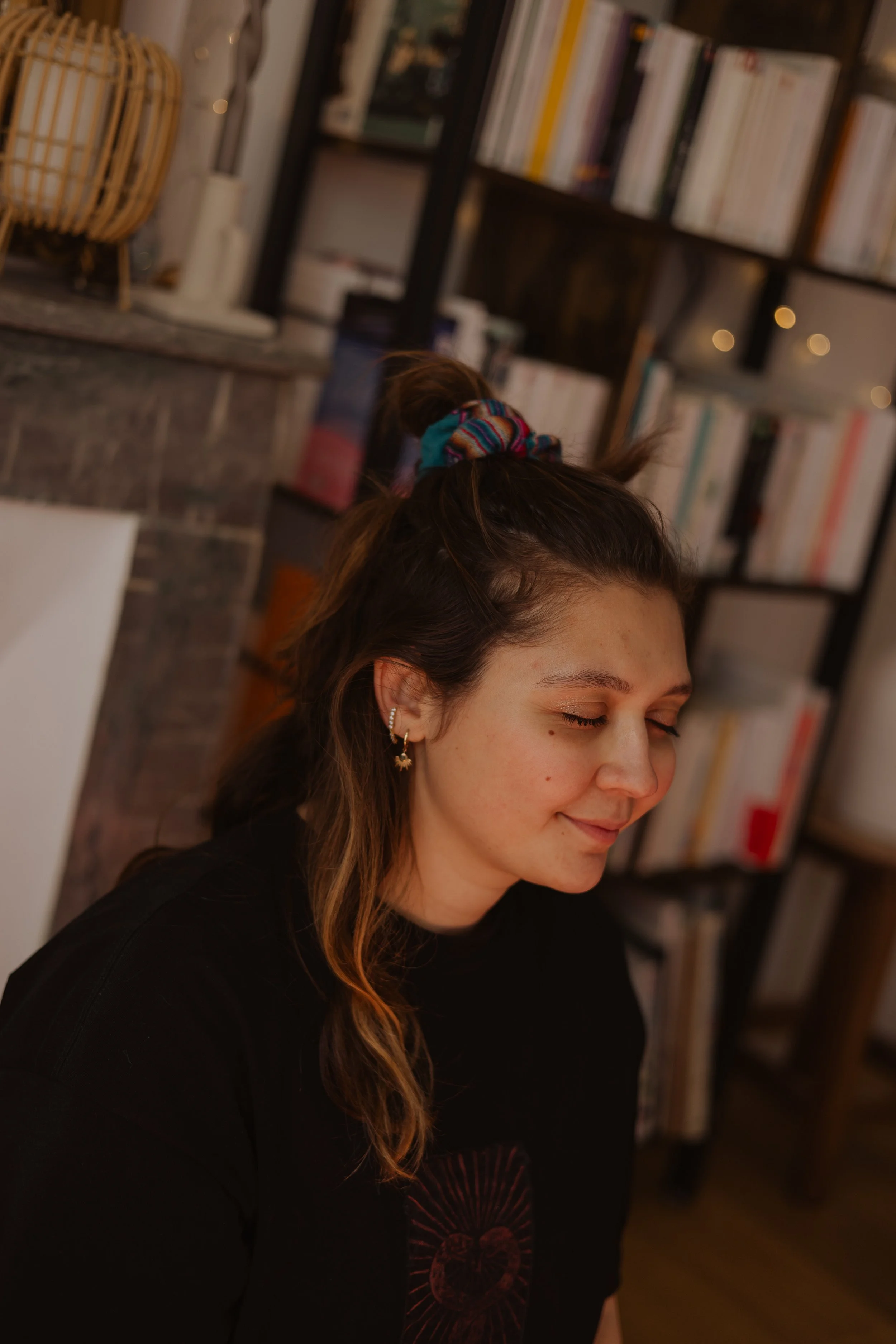 Une jeune femme avec des boucles d'oreilles, les yeux fermés, souriant légèrement, qui médite, avec une bibliothèque en arrière-plan.