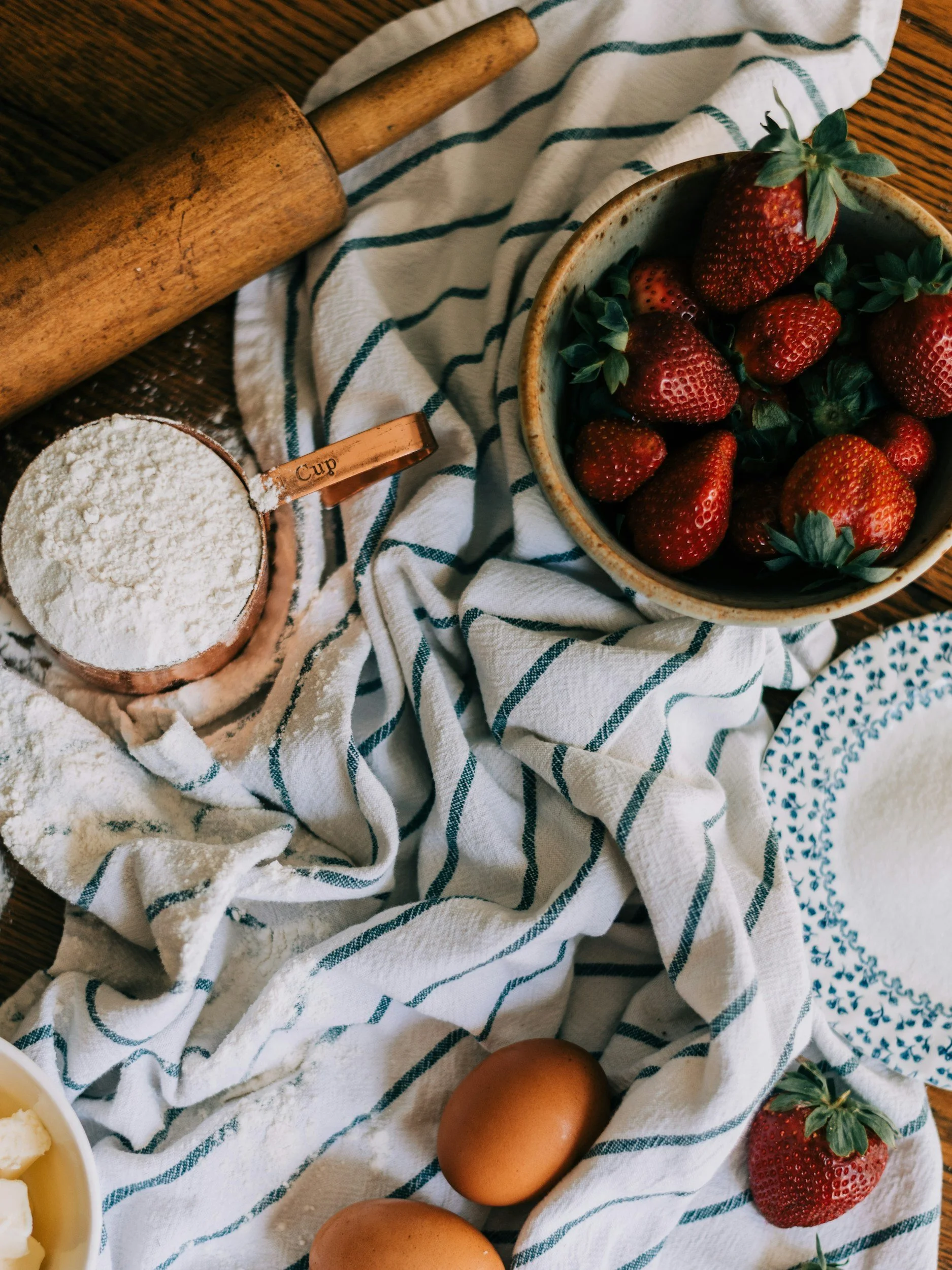 Fresh strawberries, eggs, and flour on a kitchen table with a rolling pin and a striped cloth.