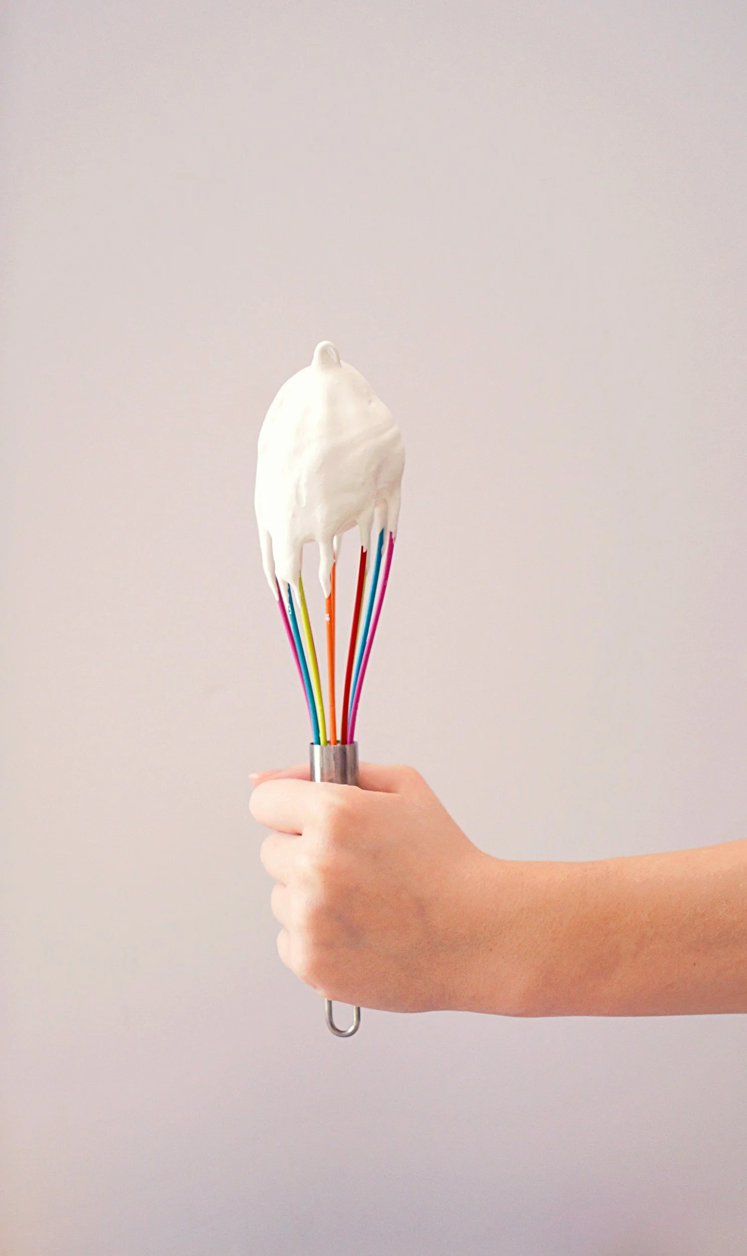 A person's hand holding a wire whisk with colorful rainbow wires, topped with white whipped cream.