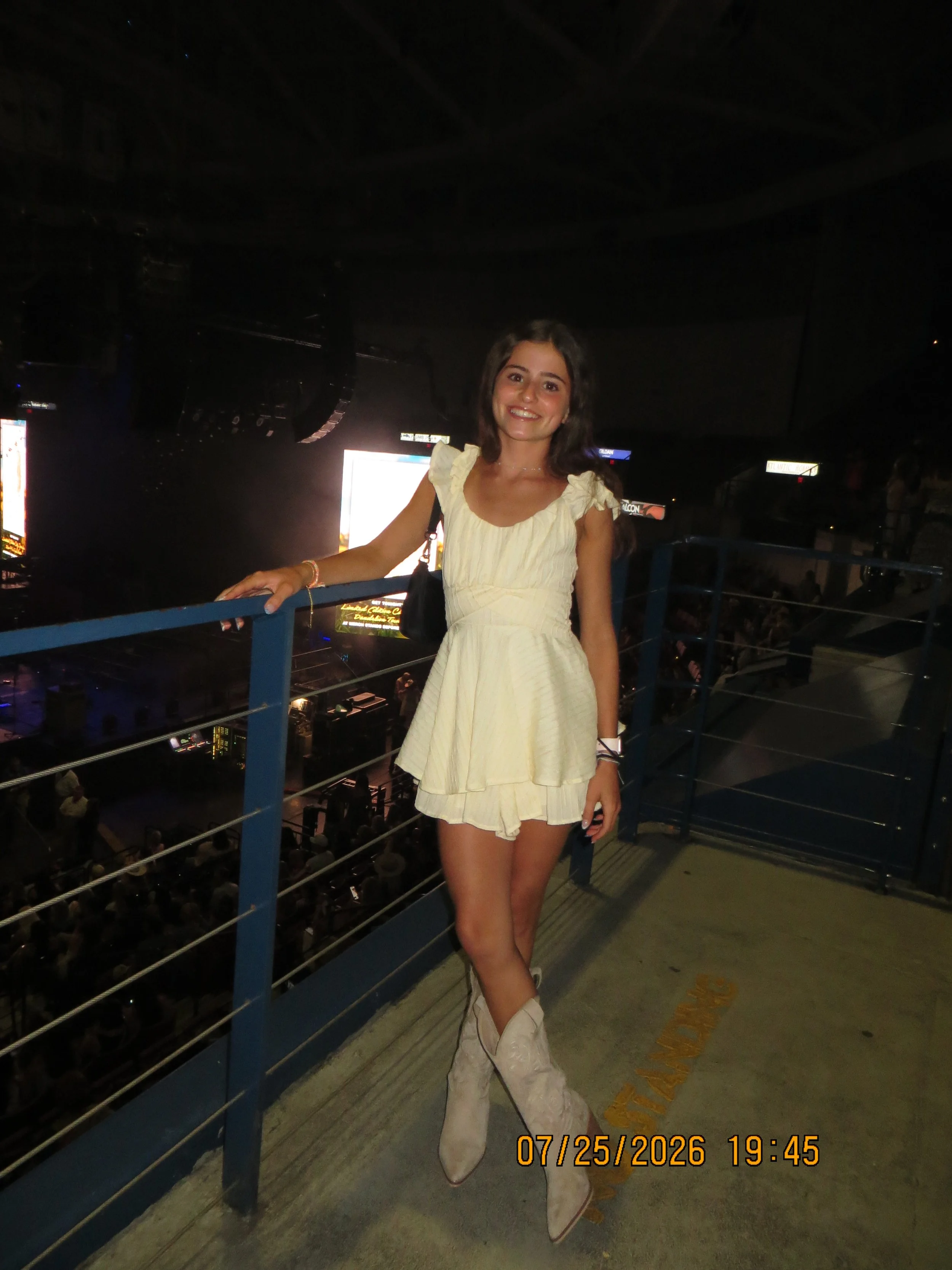 A young woman in a yellow dress and cowboy boots posing on a balcony at night with a dark indoor arena below and illuminated advertisements in the background.