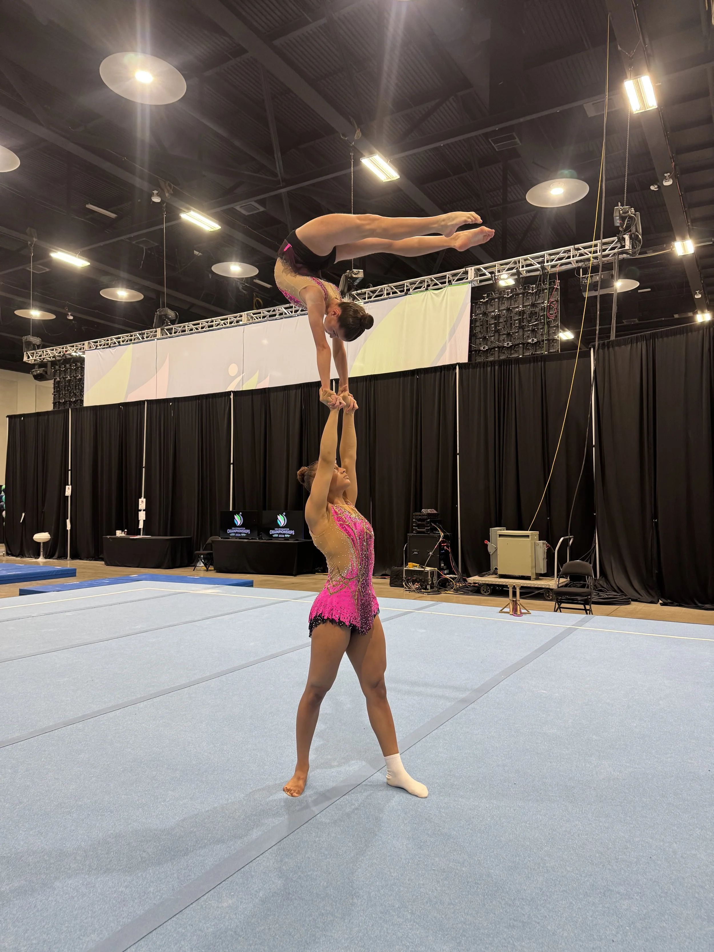 A gymnast performs a balanced handstand on the shoulders of another gymnast during a gymnastics routine in a competition setting.