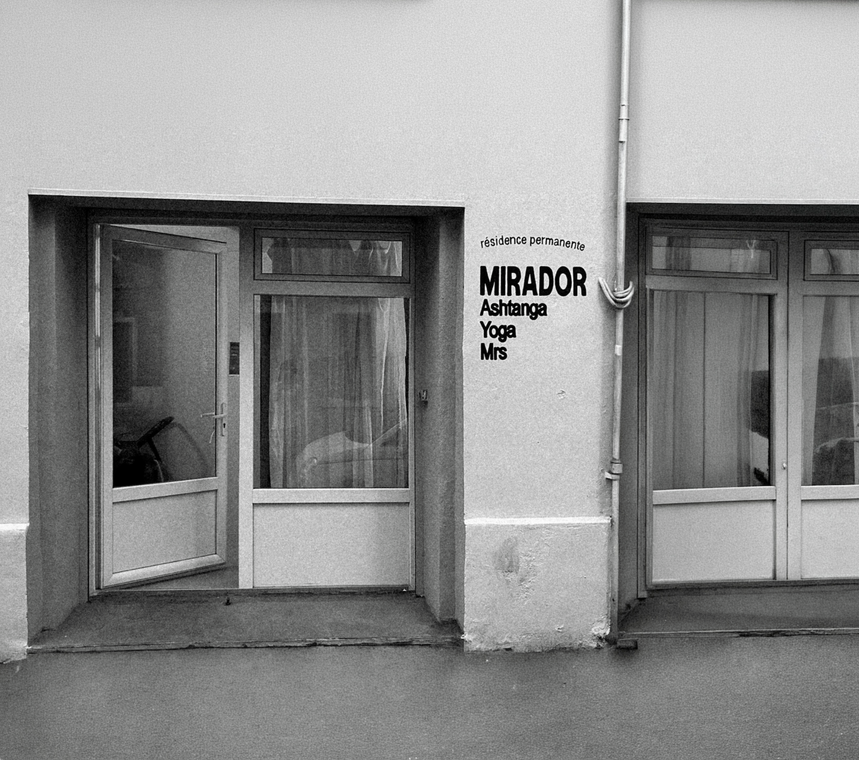 Façade d'un bâtiment blanc avec deux fenêtres et une porte en verre. À côté de la porte, il y a une inscription en français indiquant une résidence permanente avec les activités suivantes : Mirador, Ashtanga, Yoga, Mrs.