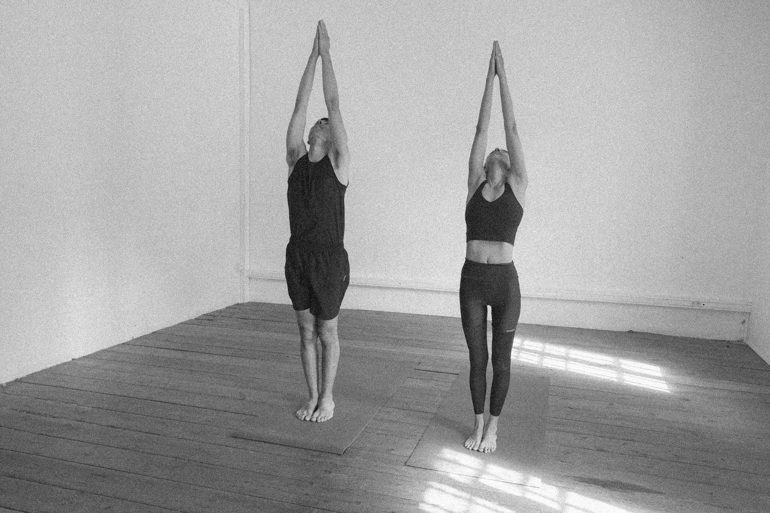 Deux femmes pratiquent le yoga dans une salle en bois, debout les bras tendus vers le haut avec les paumes jointes.