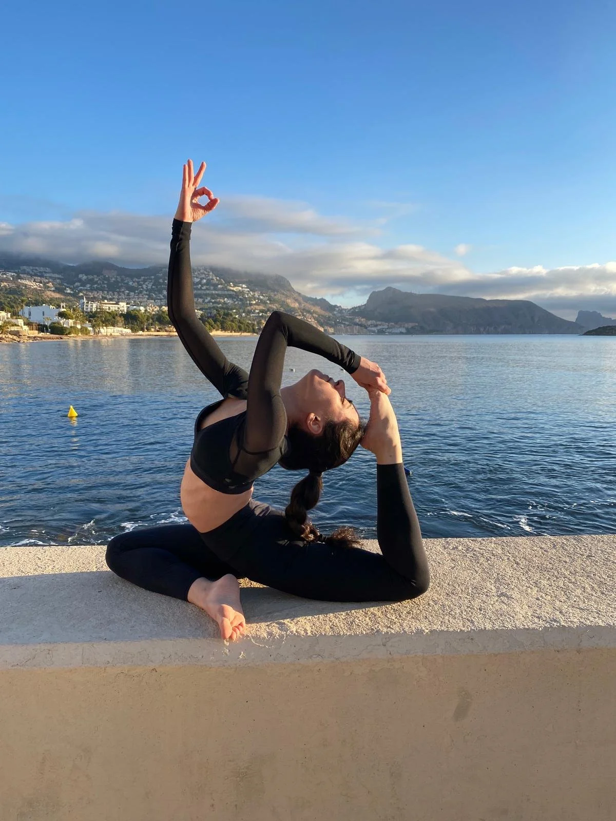 Mujer haciendo yoga en un poyete la orilla del mar en un día soleado con montañas en el fondo.
