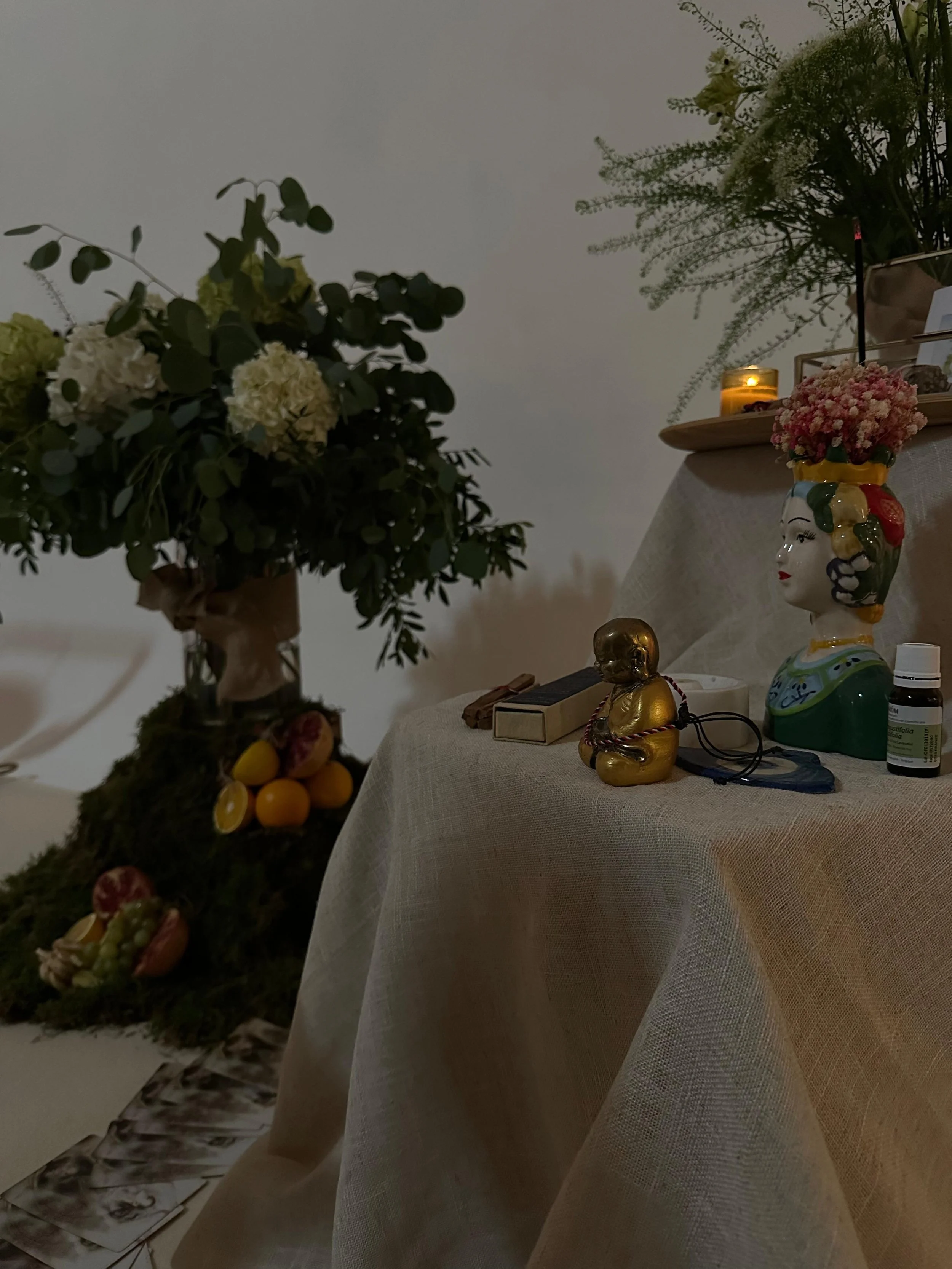 Imagen de un altar con flores, una vela encendida, figuras decorativas, y frutas en la base, en un ambiente con paredes blancas.