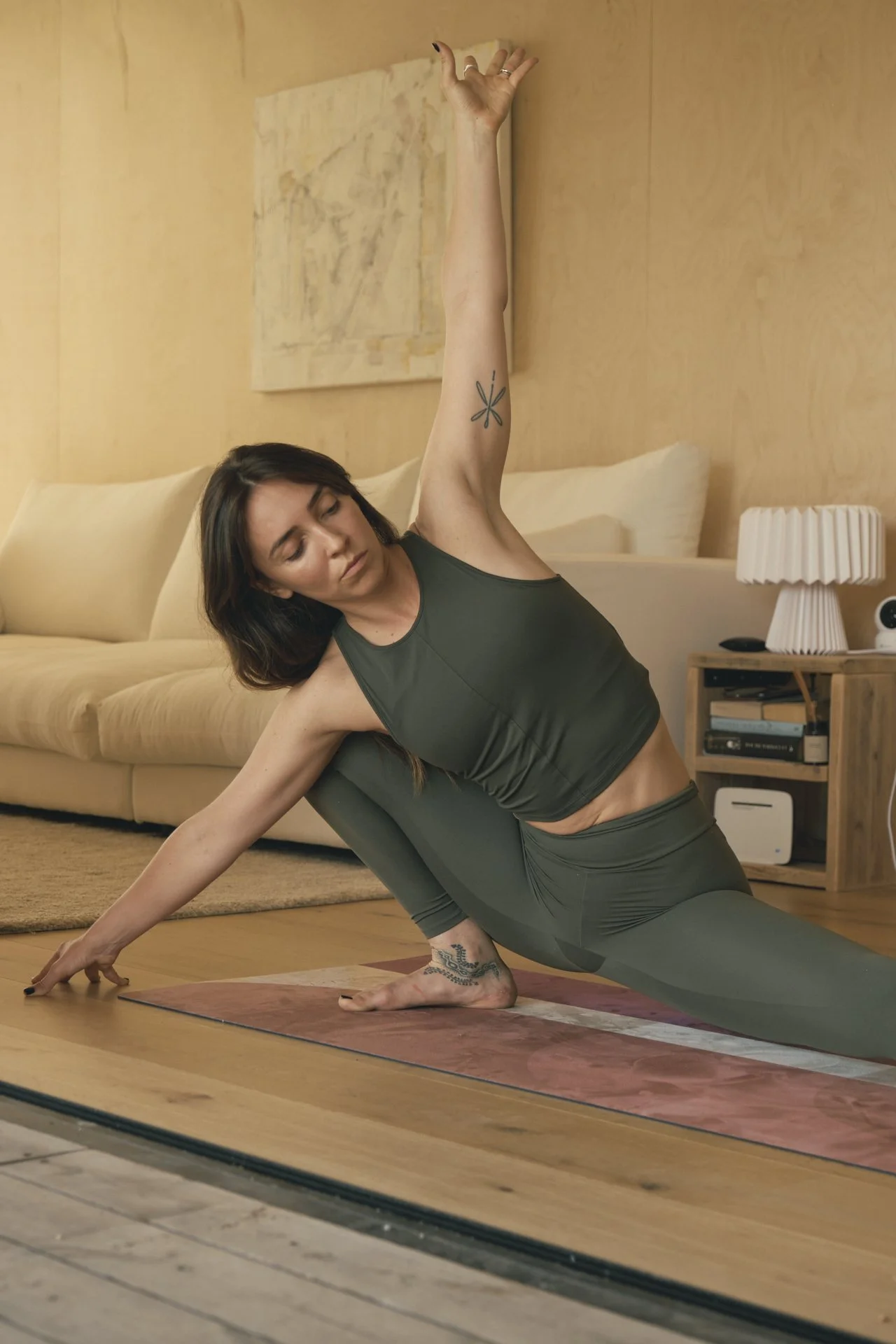 Una mujer practicando yoga en un tapete en una sala de estar, realizando una postura lateral con mano levantada y expresión concentrada.