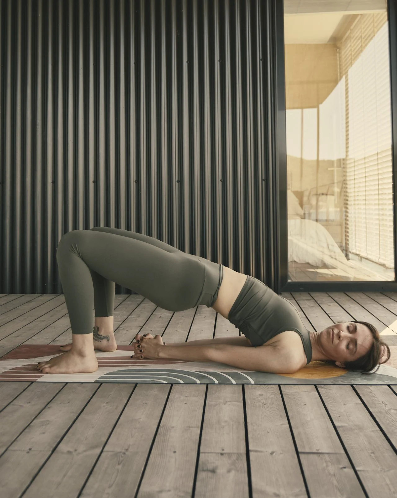 Una mujer practicando yoga en un espacio con piso de madera y paredes de metal y vidrio, realizando una postura de puente en una colchoneta con fondo de cama y ventanas con persianas.