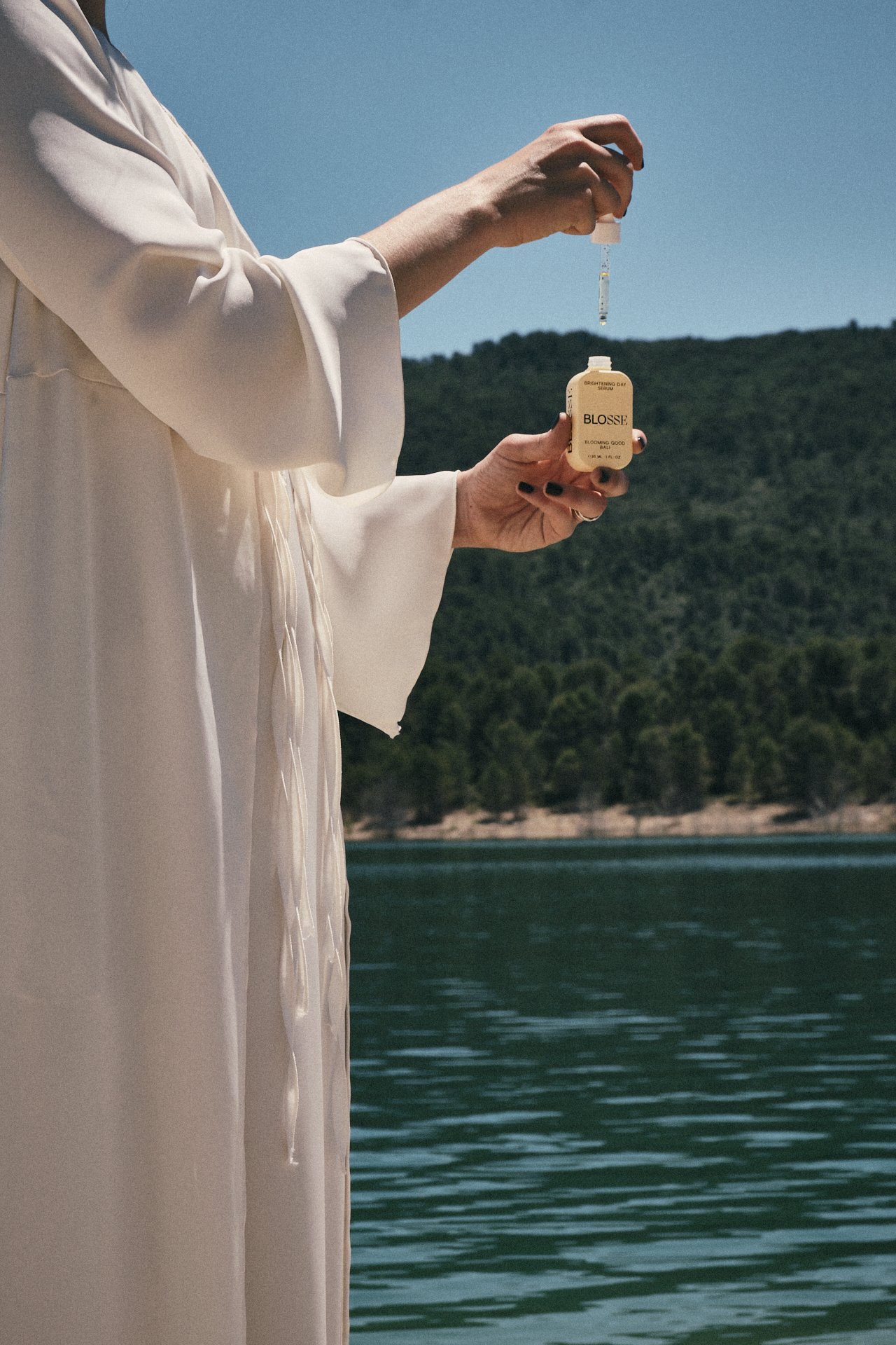 Persona sosteniendo un frasco de cosmética líquida al aire libre cerca de un cuerpo de agua con colinas verdes al fondo, en un día soleado.