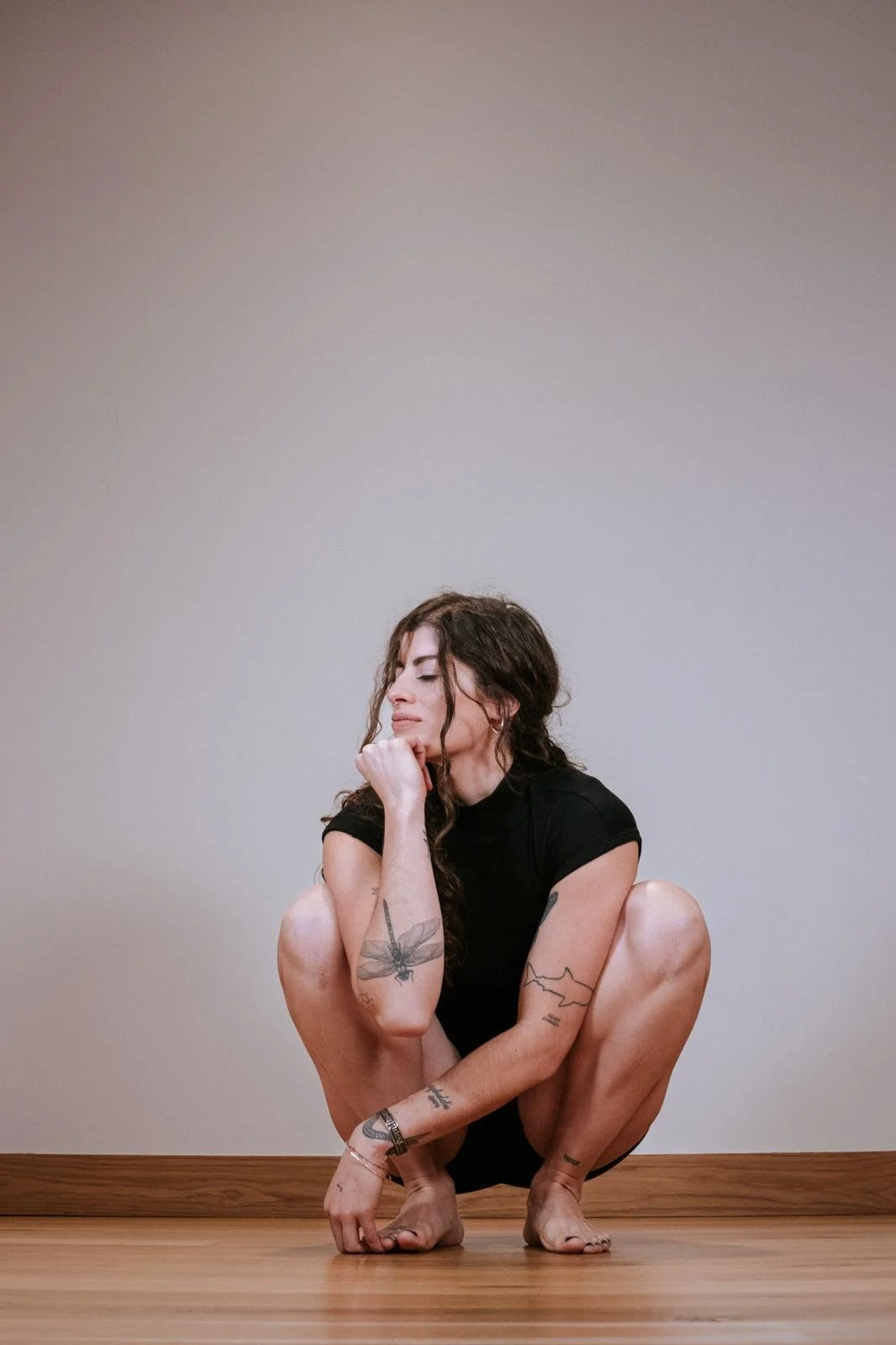 Mujer con tatuajes en una postura de yoga, en un espacio interior con suelo de madera y pared gris.