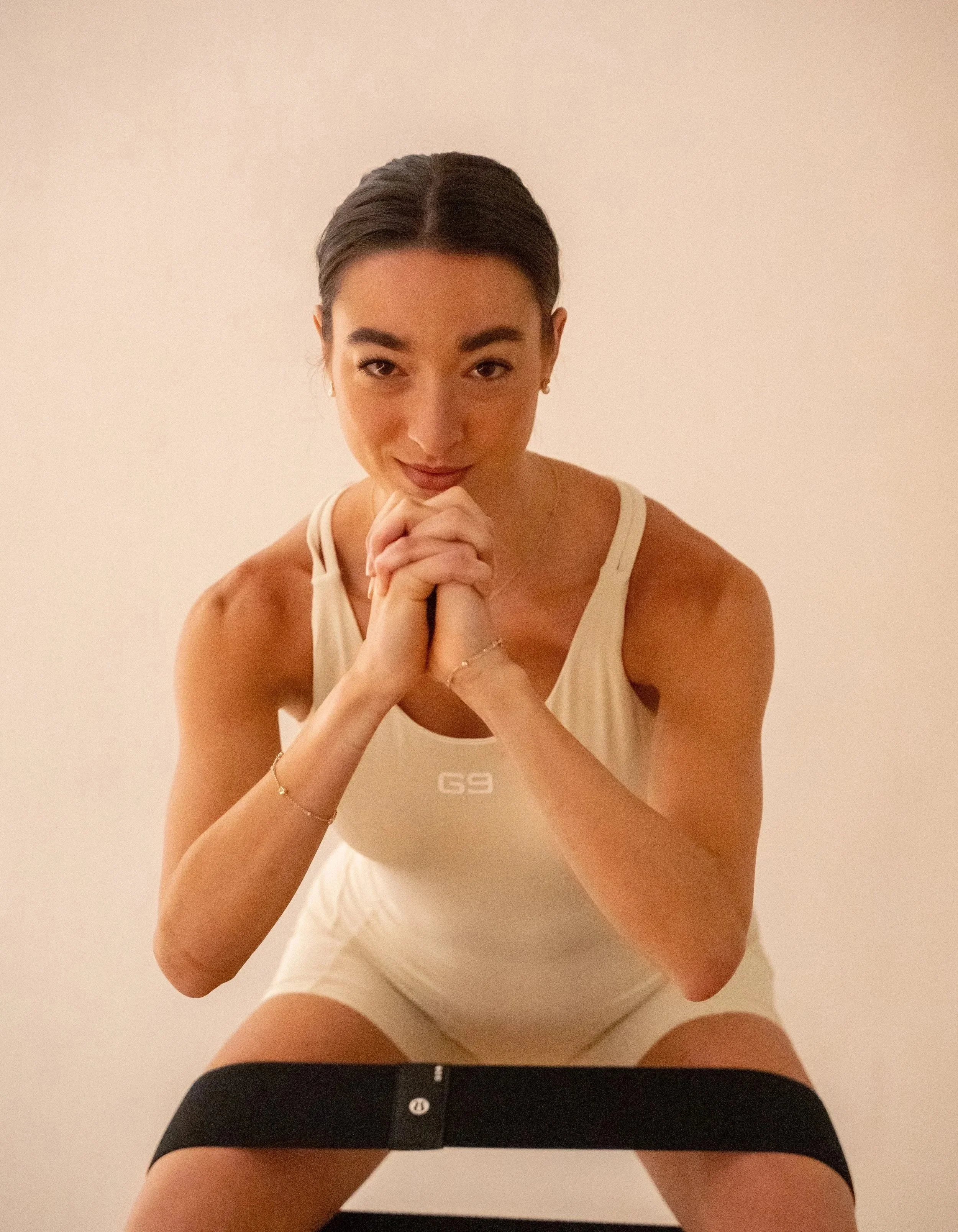 Mujer con cabello oscuro en una coleta, posando con manos juntas bajo el rostro, usando ropa clara y accesorios sencillos, en un fondo neutro.