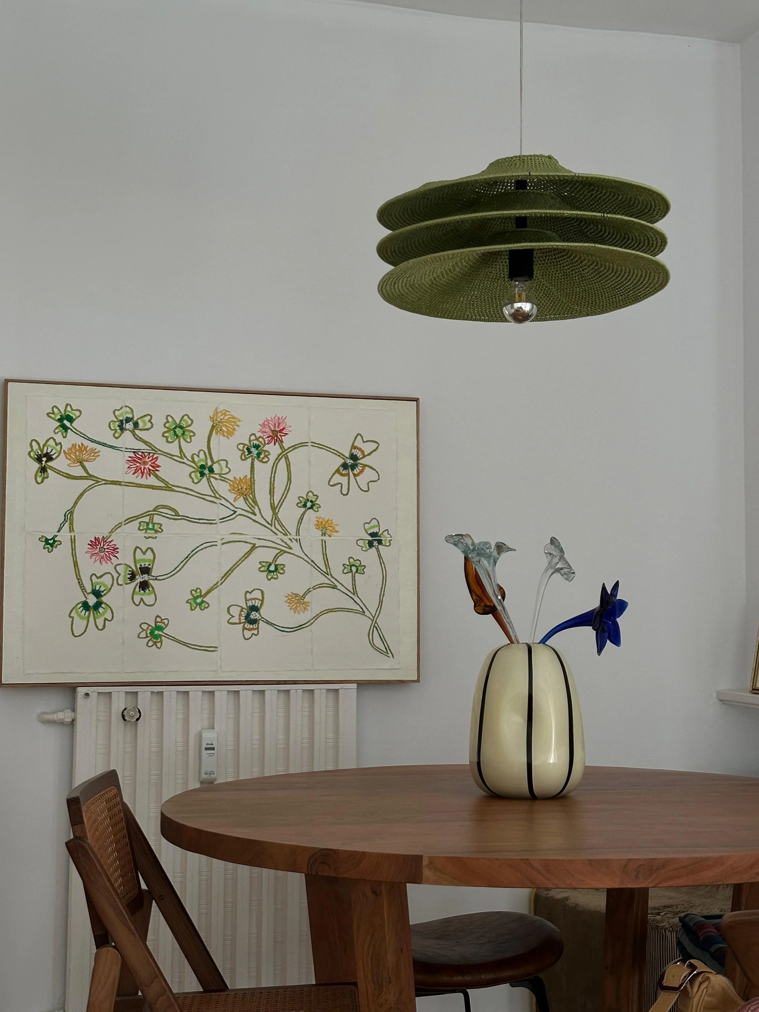 Interior de una sala con mesa de madera, una lámpara decorativa moderna en el centro, un cuadro con diseño floral en la pared y una base de radiador debajo de este. También hay una silla visible a la izquierda y objetos decorativos, como figuras de vidrio con forma de flores y un jarrón con arreglos florales.