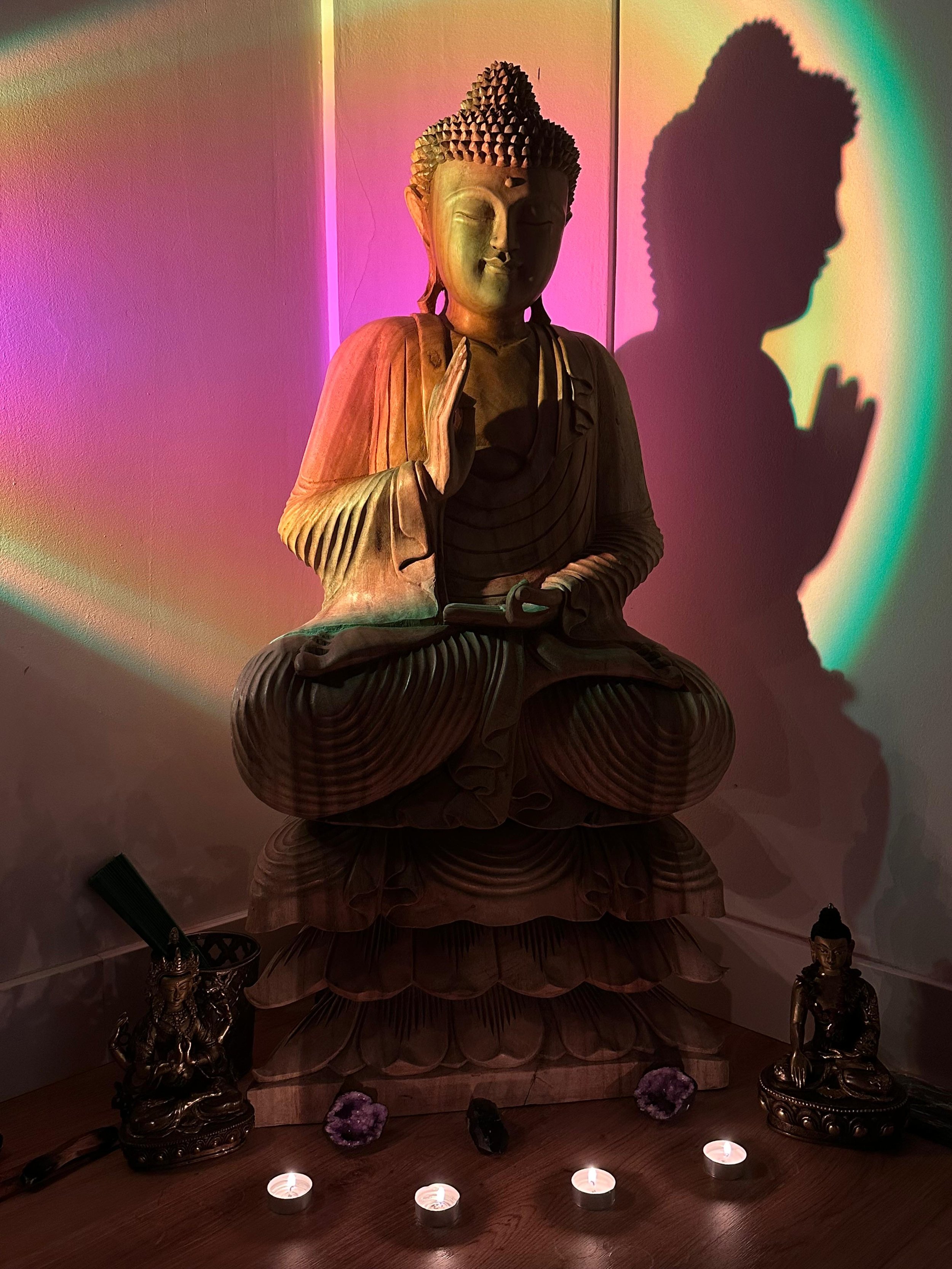 Escultura de Buda de madera, iluminada con luces de colores suaves, rodeada de pequeñas velas y figuras decorativas, creando un ambiente de tranquilidad y meditación en un espacio interior.