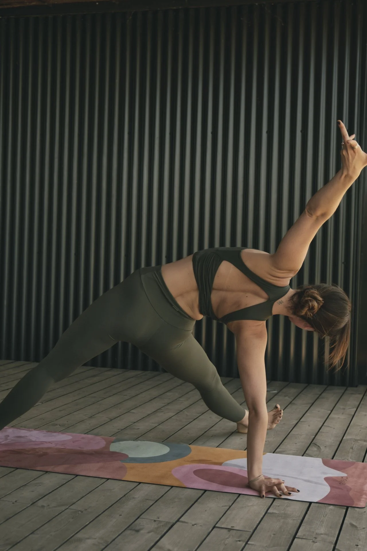 Mujer haciendo yoga en una postura de ángulo lateral con una mano en el suelo y la otra levantada, en un deck de madera frente a una pared con paneles oscuros.