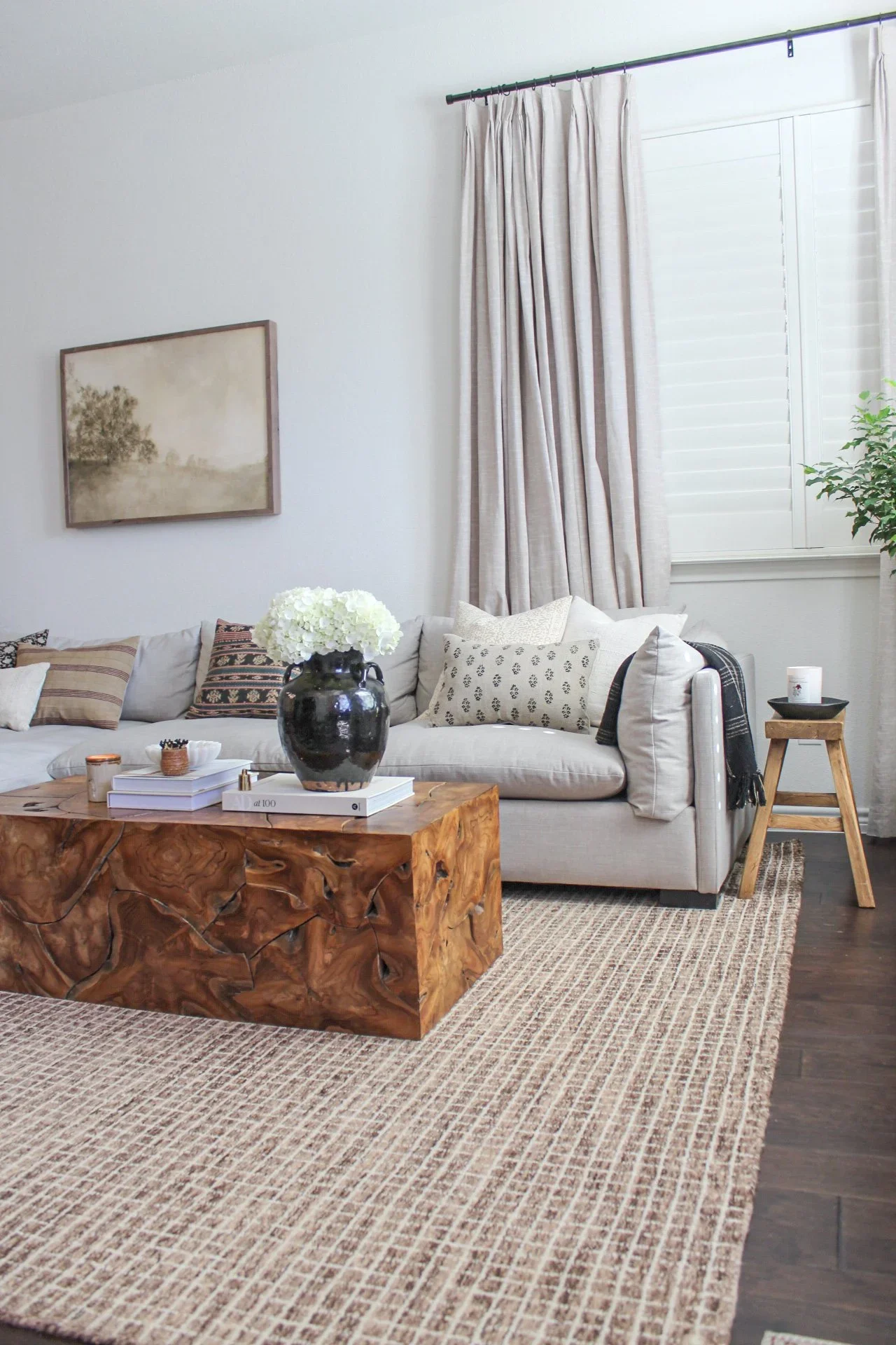 A cozy living room with a white sofa, patterned pillows, a wooden coffee table with a black vase of white flowers, a framed landscape painting, and light-colored curtains by a window. There is a small wooden side table with a black bowl and candle.