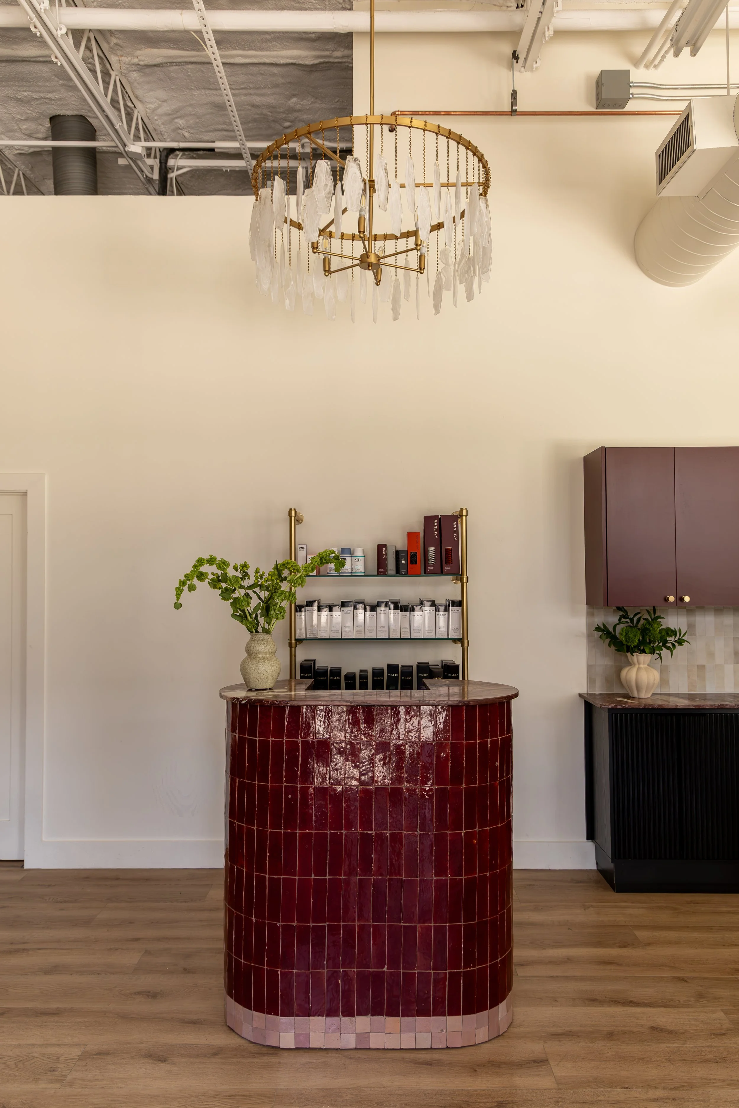 A reception area featuring a red tiled curved counter, two potted plants on either side, wall shelves with products, and a gold chandelier hanging from an exposed ceiling with industrial pipes.
