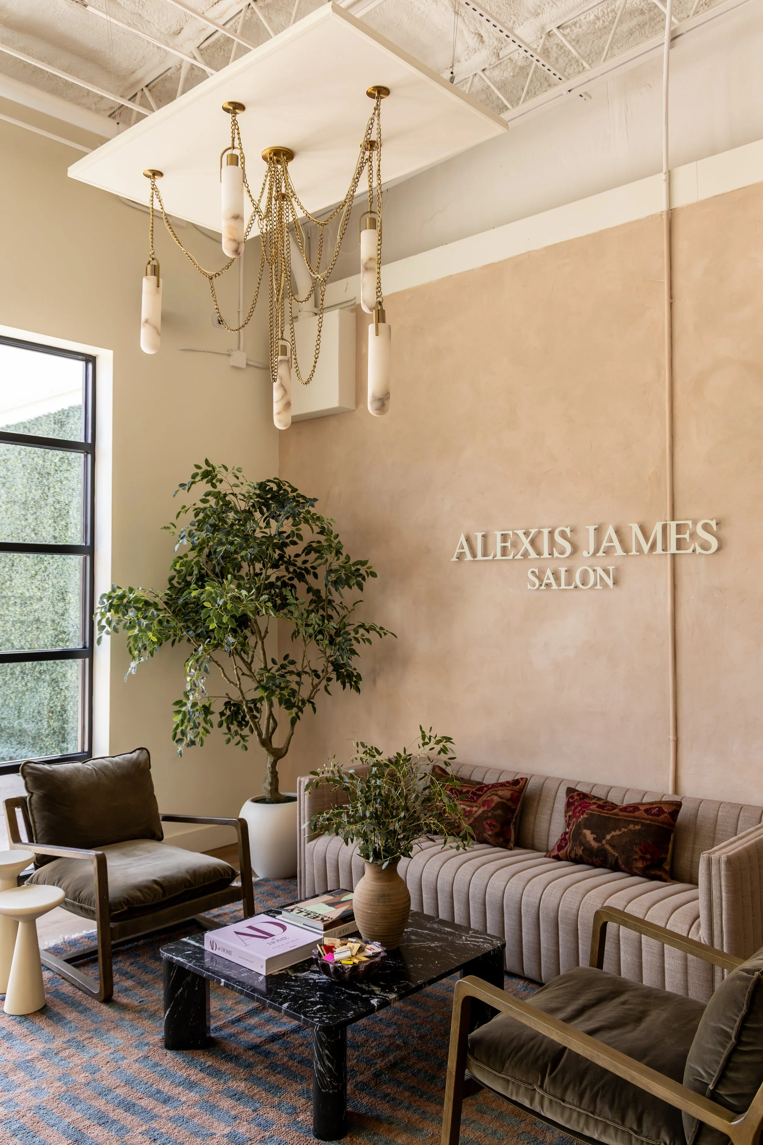 Interior of a salon with seating area, potted plants, a black marble coffee table, books, and a modern chandelier hanging from the ceiling. The salon's name 'ALEXIS JAMES SALON' is displayed on the wall.