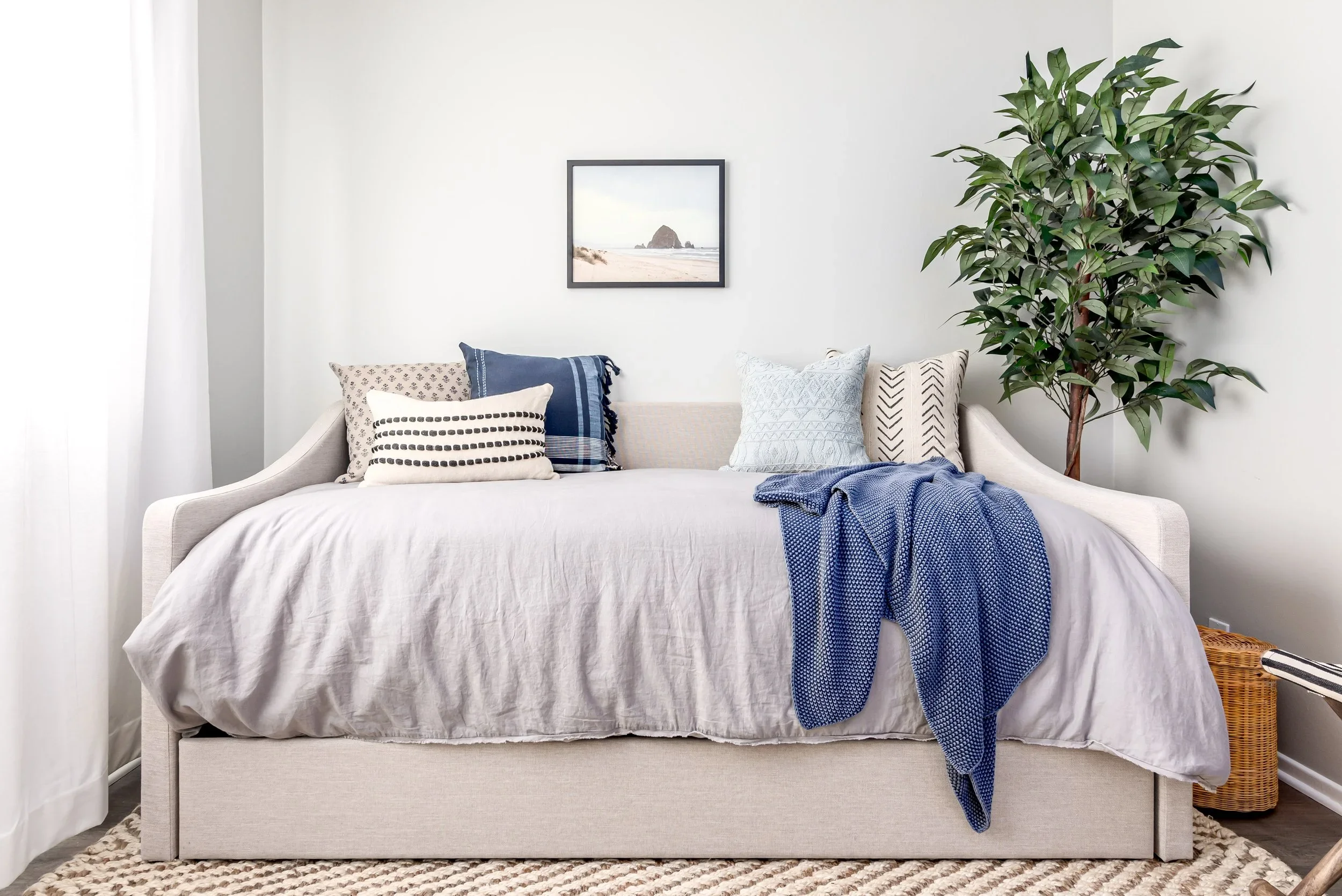 A bedroom with a bed covered in a white bedspread, decorated with patterned and solid pillows, and a blue throw blanket. There is a large green leafy plant in a basket on the right and a framed beach scene photograph on the wall.