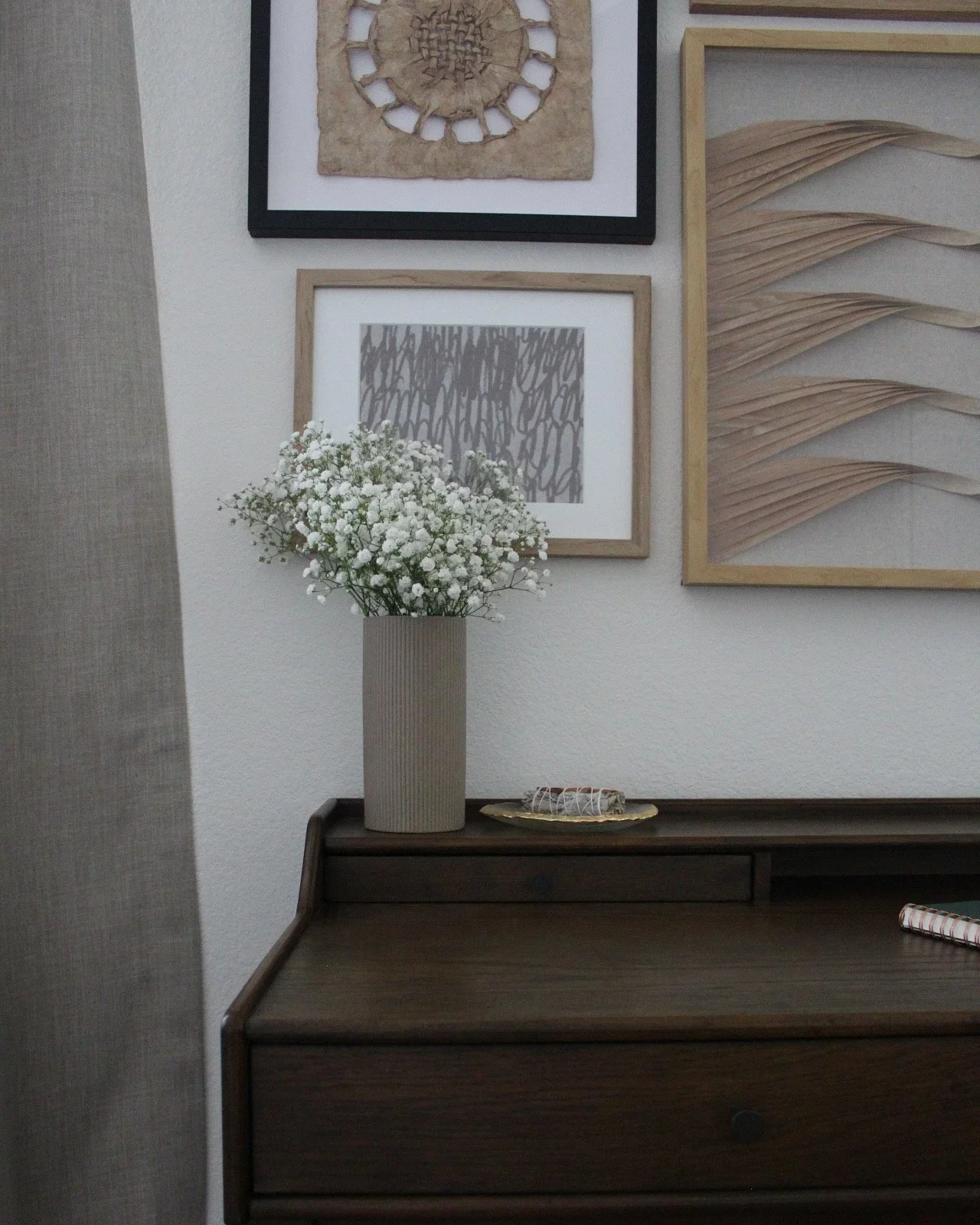 A dark wooden desk with a beige ribbed ceramic vase filled with small white flowers. Behind the vase is a gold-trimmed ceramic plate with decorative twine. On the wall above are framed art pieces including a black and white abstract print, a textured